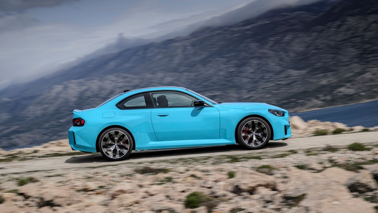 Carro esportivo azul-claro, modelo cupê, em movimento numa estrada de terra, com montanhas escuras e um lago azul ao fundo