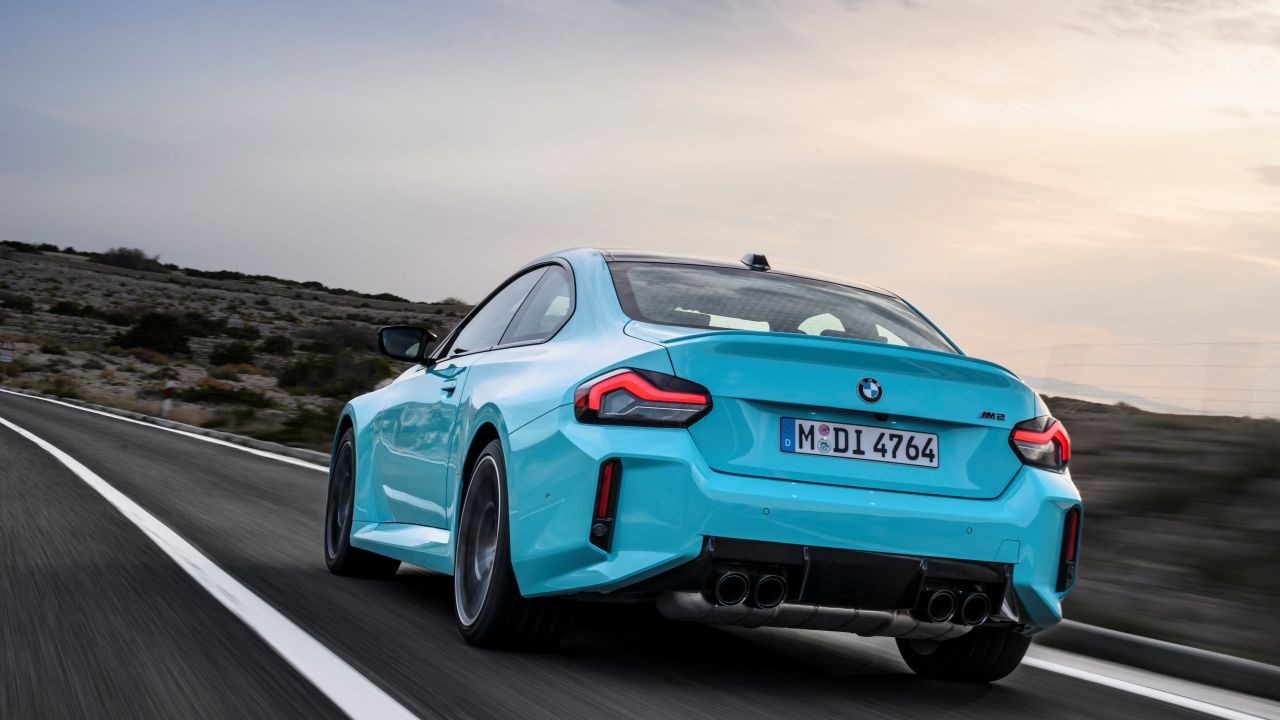 Carro esportivo BMW M2 azul-turquesa visto por trás, acelerando em uma estrada sinuosa com montanhas áridas ao fundo e céu nublado ao entardecer