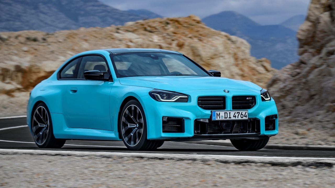 Carro esportivo BMW M2 azul-turquesa, com teto preto, parado em uma estrada de asfalto. Ao fundo, montanhas rochosas e céu azul claro. A placa do carro é M DI 4764