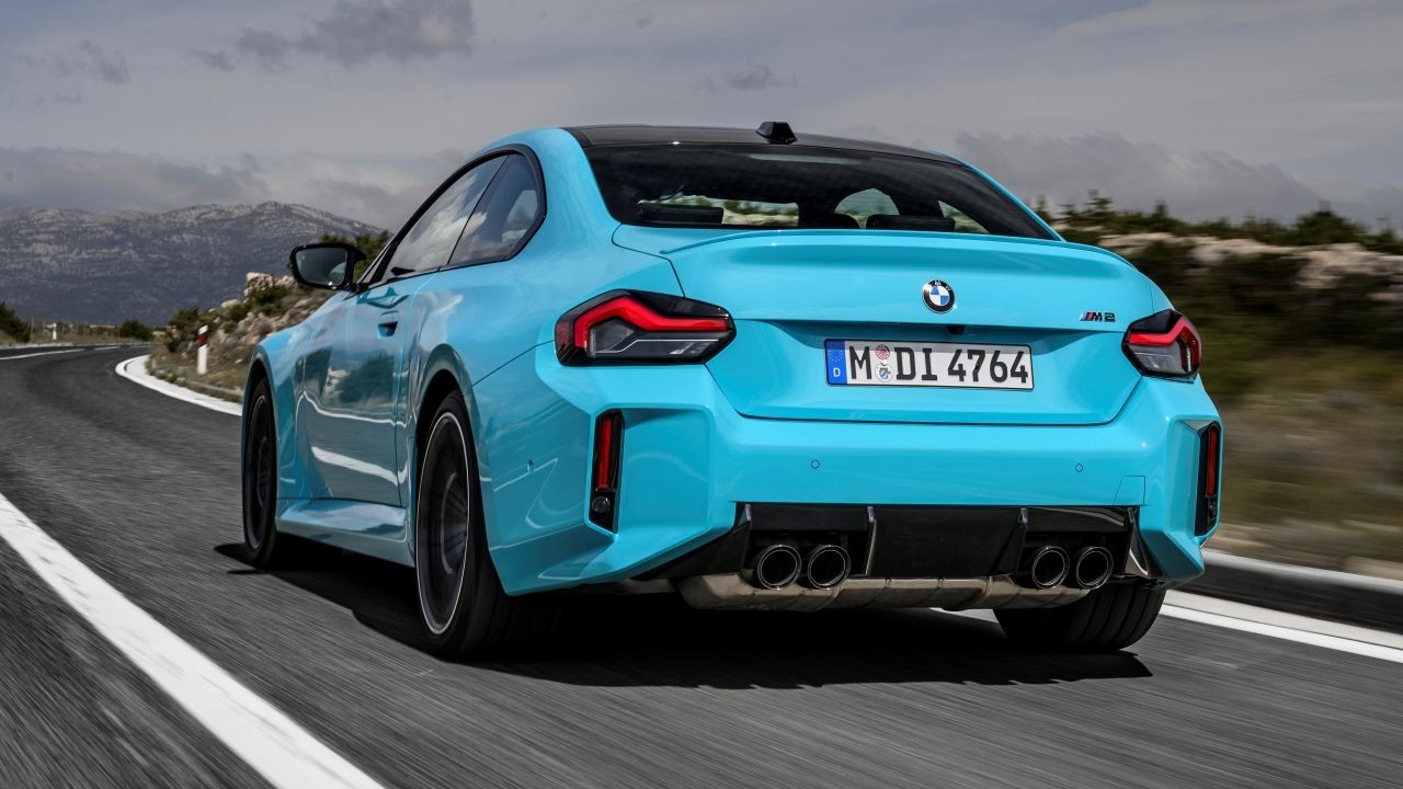 Carro esportivo BMW M2 azul-claro, visto por trás, em uma curva de estrada montanhosa. O veículo tem quatro saídas de escapamento e lanternas traseiras escuras, com montanhas e céu nublado ao fundo