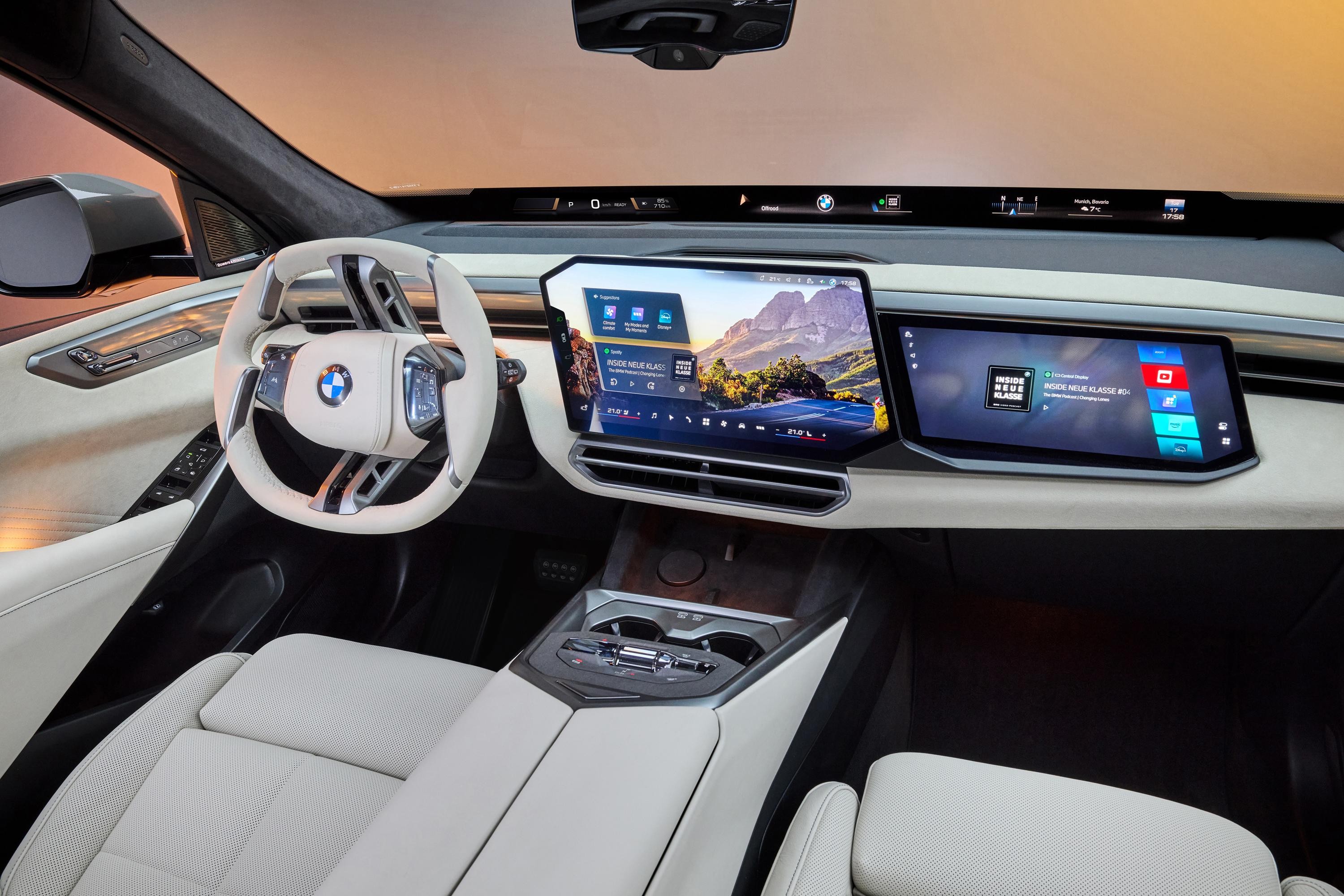 Interior de um carro moderno com volante branco e painel digital. Duas telas grandes exibem um mapa com montanhas e um menu de aplicativos. Bancos claros e console central com porta-copos. O para-brisa reflete um céu alaranjado
