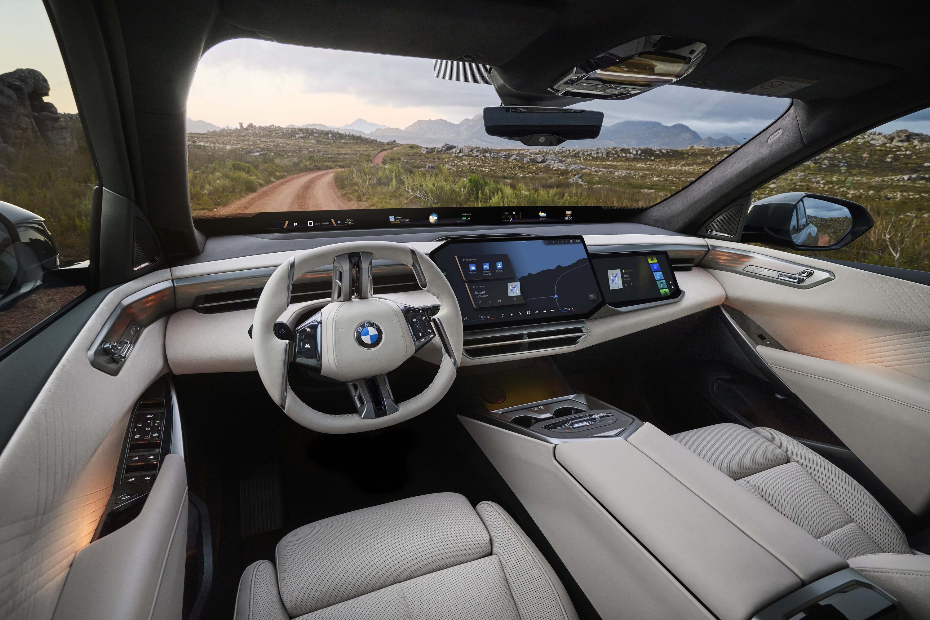 Interior de um carro BMW moderno, com volante branco e o painel digital exibindo um mapa. Ao fundo, uma estrada de terra serpenteia por uma paisagem montanhosa e verdejante sob um céu parcialmente nublado