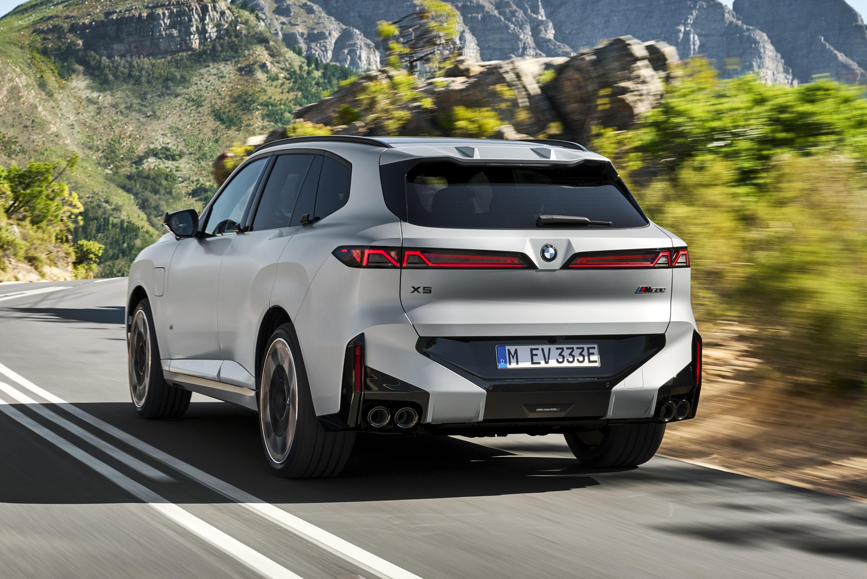 SUV BMW X5 cinza claro, visto por trás, em uma estrada sinuosa com montanhas e vegetação ao fundo