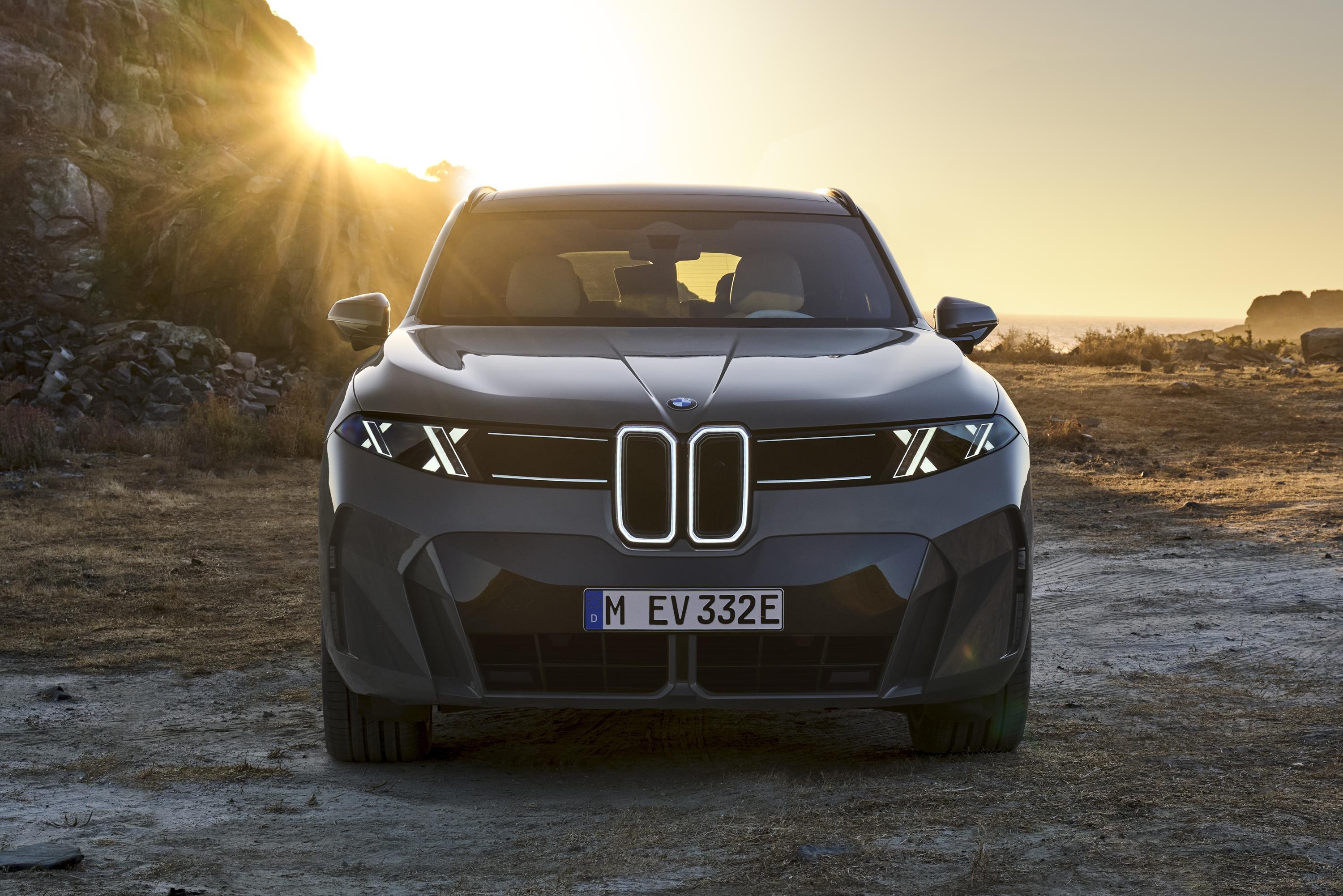 Carro BMW elétrico cinza escuro, visto de frente, com faróis em X e grade iluminada, estacionado em terreno árido ao pôr do sol