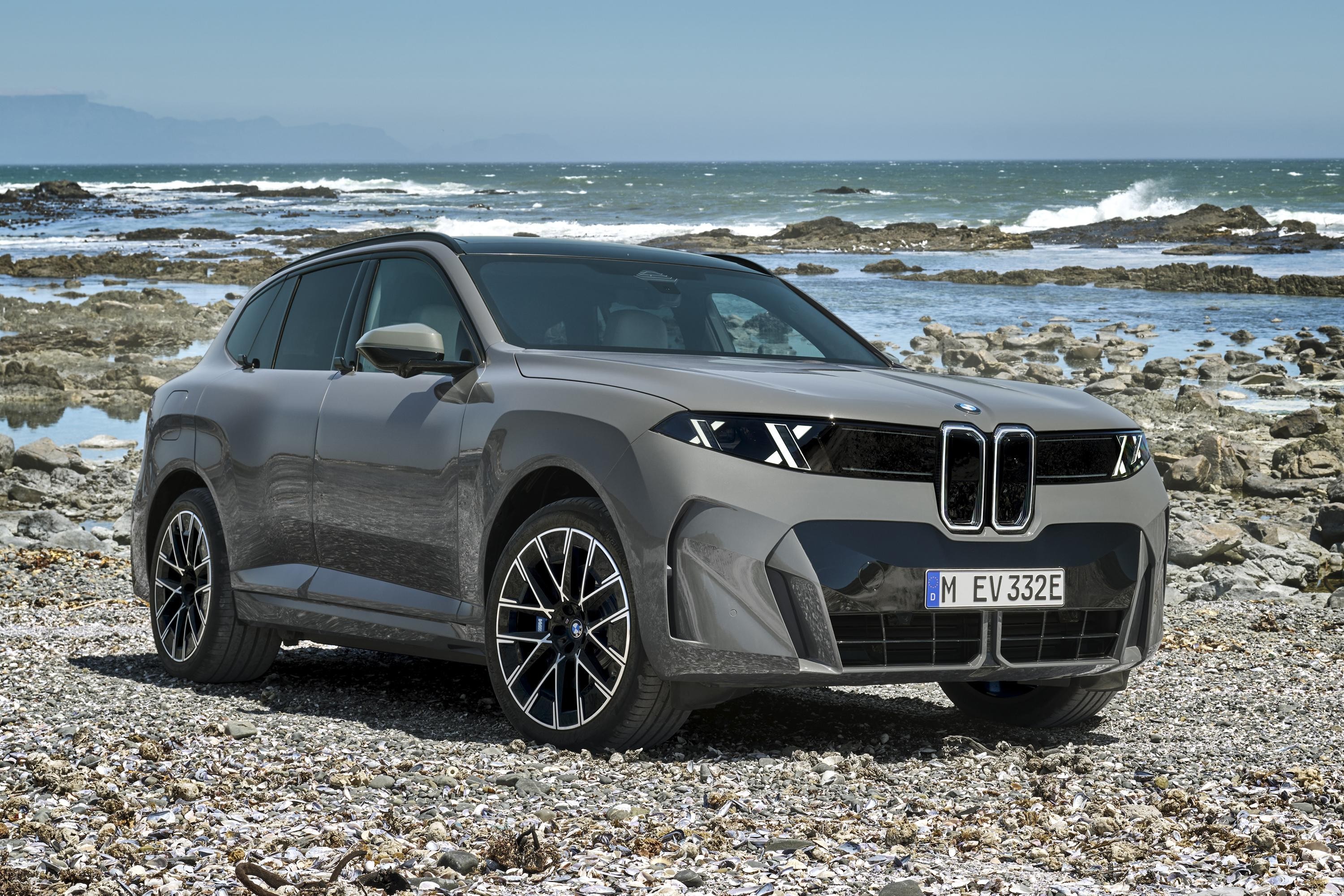 SUV BMW cinza-chumbo estacionado em uma praia rochosa, com o mar azul e montanhas ao fundo. O carro tem faróis em X e grade frontal dupla preta, com a placa M EV 332E. O céu está claro e o sol brilha