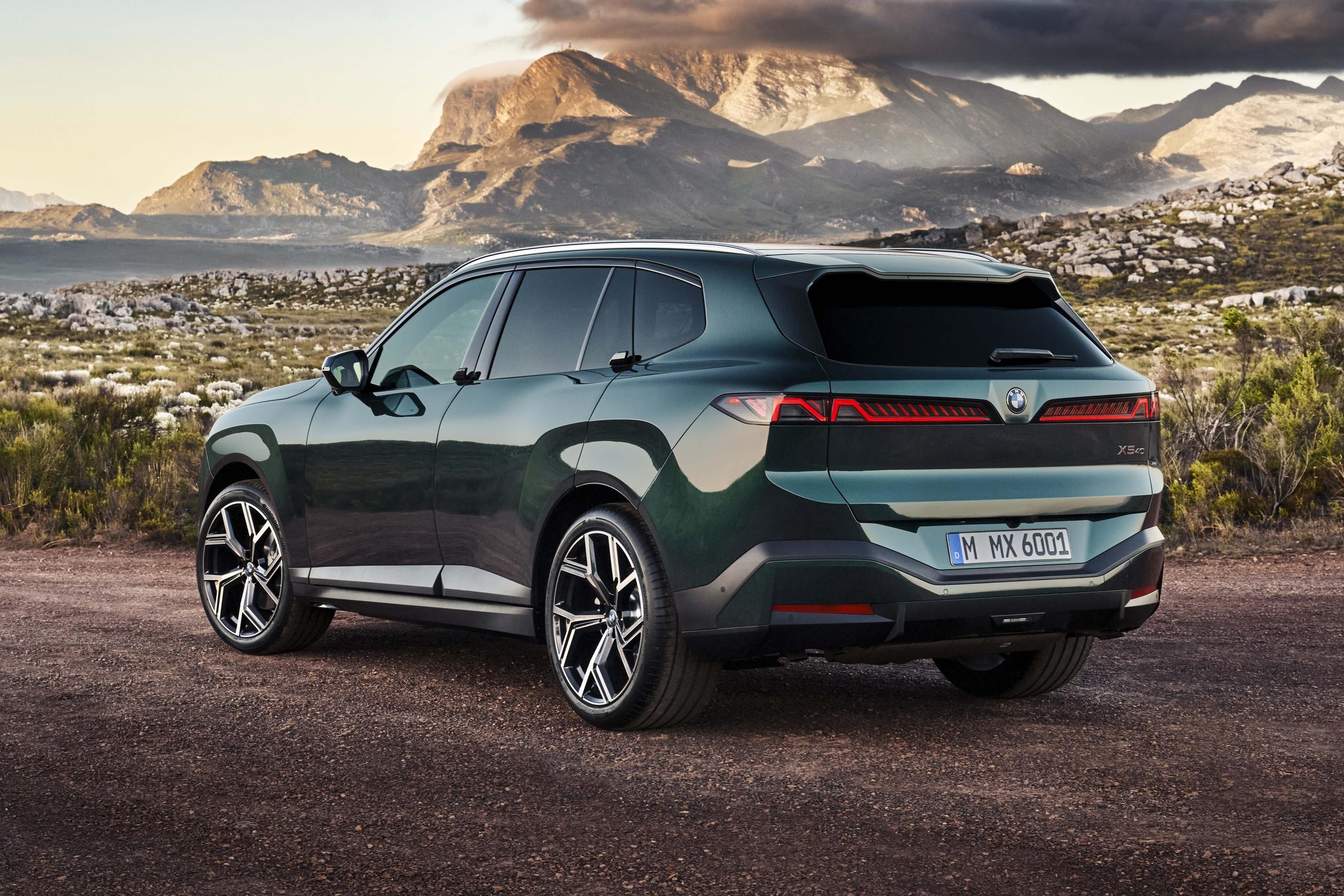 SUV BMW XM verde-escuro, visto de trás e lateral, estacionado em estrada de terra com montanhas e céu nublado ao fundo
