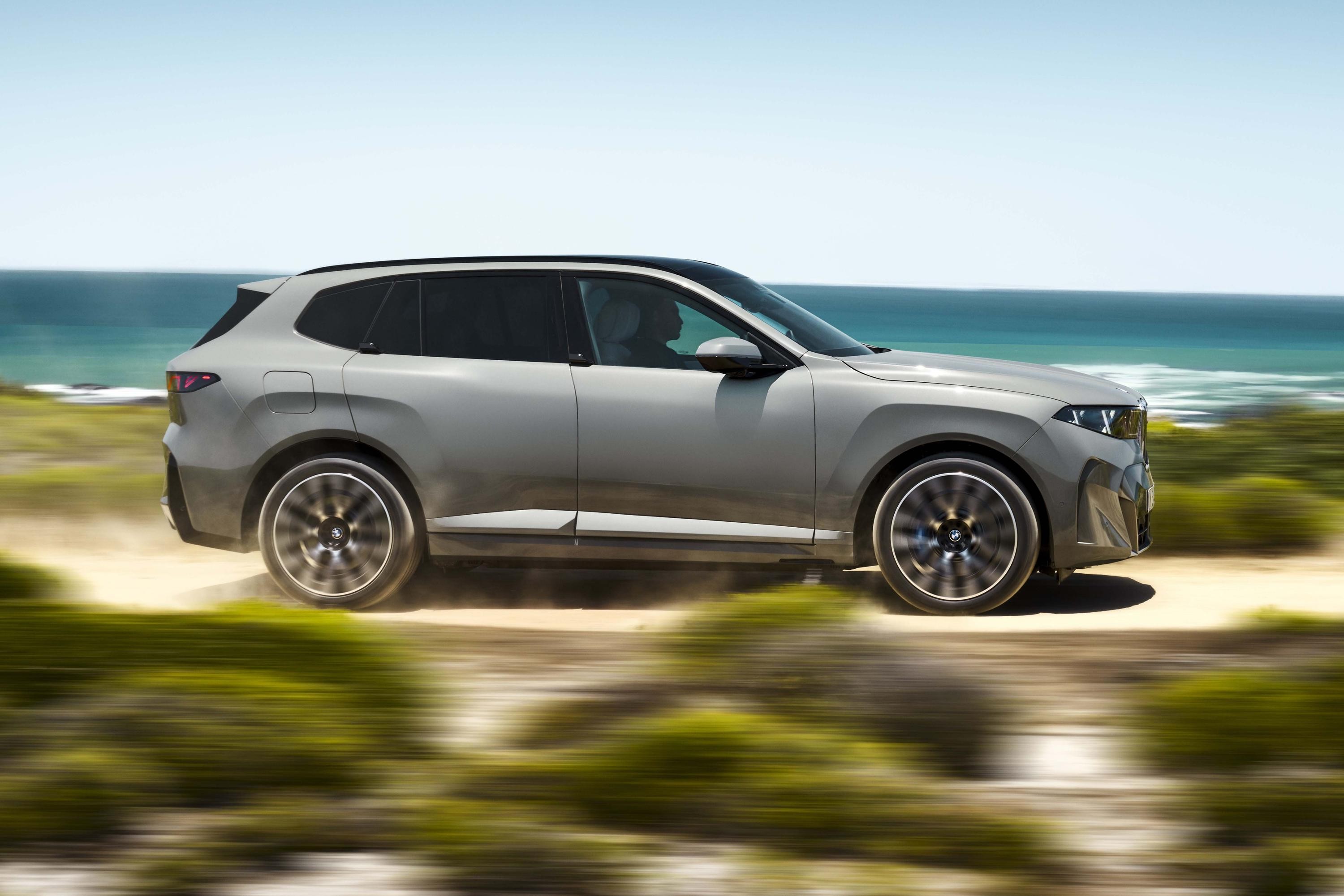 SUV cinza-claro em movimento, visto de lado, com rodas girando rapidamente em uma estrada de terra. Ao fundo, o oceano azul e o céu claro, com vegetação borrada em primeiro plano, indicando velocidade