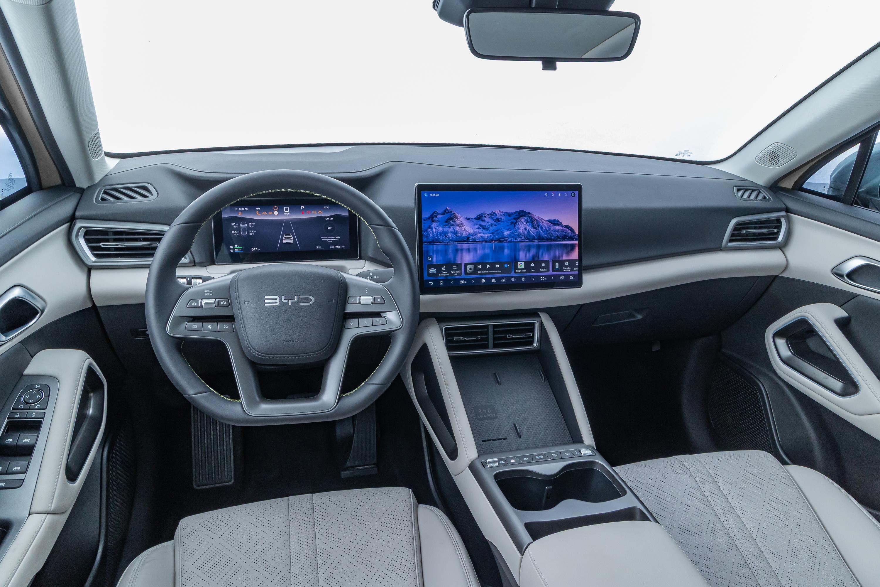 Interior de um carro moderno com volante multifuncional exibindo o logo BYD. O painel possui duas telas digitais: uma menor atrás do volante com informações de navegação e uma maior central mostrando uma paisagem de montanhas nevadas. Os bancos são cinza claro com detalhes em losango, e o console central tem porta-copos e botões de controle. O acabamento interno combina tons de cinza, preto e branco, com detalhes metálicos nas maçanetas e saídas de ar