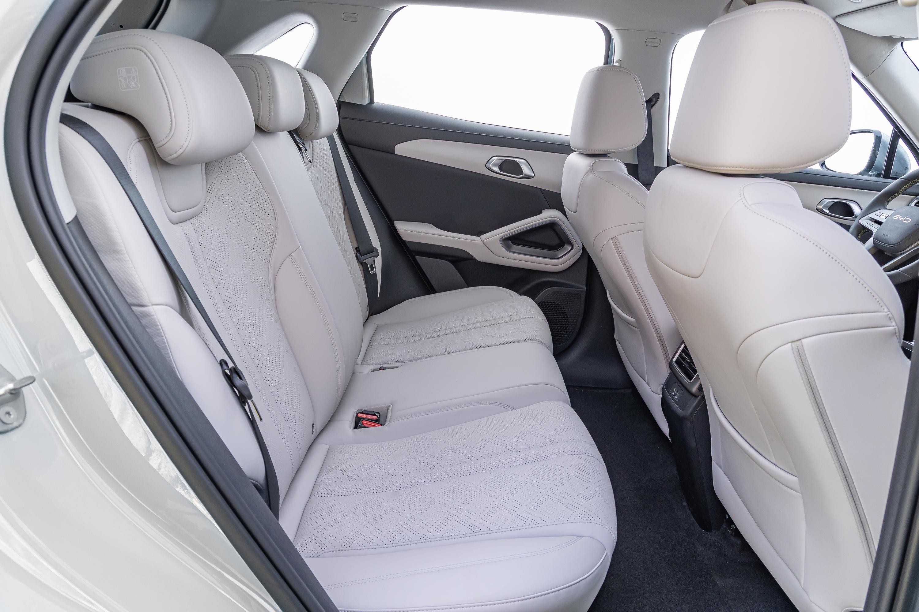 Interior de carro com bancos traseiros e dianteiros em couro branco, com detalhes em cinza escuro nas laterais e no assoalho. Os bancos traseiros possuem três encostos de cabeça e cintos de segurança pretos. A porta traseira tem acabamento em cinza escuro e branco, com maçaneta cromada. O banco do motorista é visível parcialmente à direita