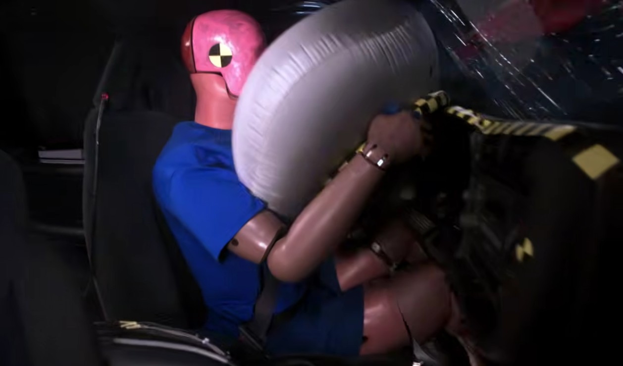 Um boneco de teste de colis&atilde;o com cabe&ccedil;a rosa e corpo azul, sentado no banco do motorista, &eacute; atingido por um airbag branco inflado durante um teste de seguran&ccedil;a veicular