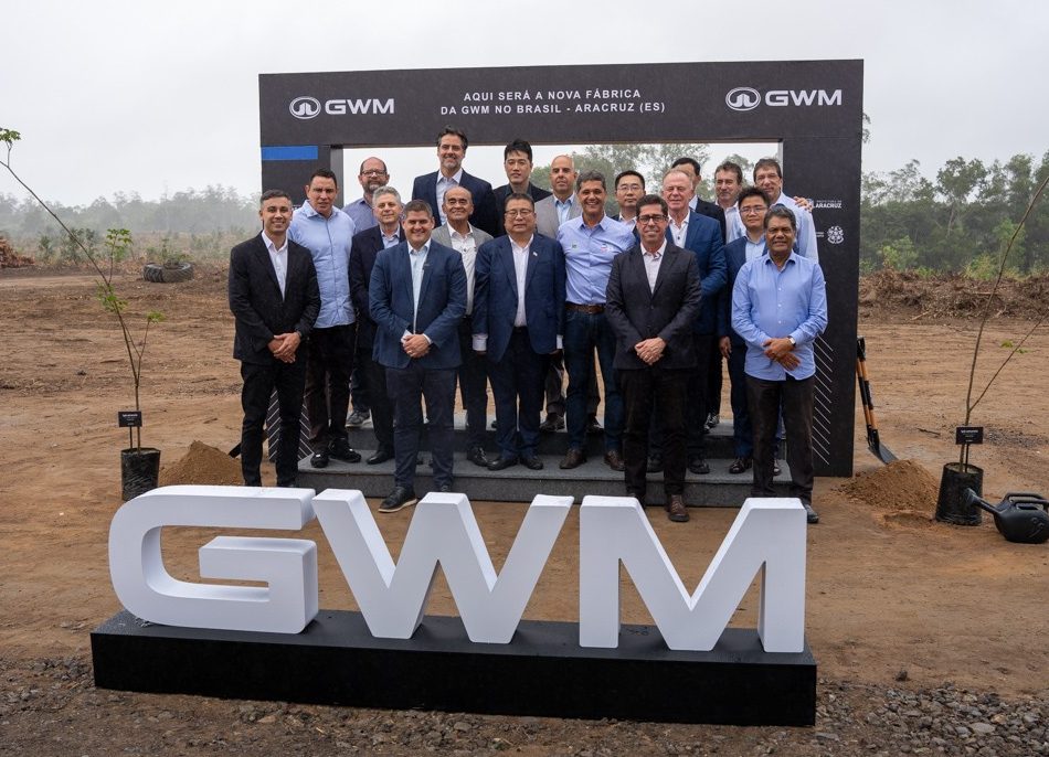 Um grupo de cerca de 20 homens, a maioria de terno, posa para foto em um terreno de terra batida. Atrás deles, um painel preto com o texto AQUI SERÁ A NOVA FÁBRICA DA GWM NO BRASIL - ARACRUZ (ES). Em primeiro plano, letras brancas formam GWM