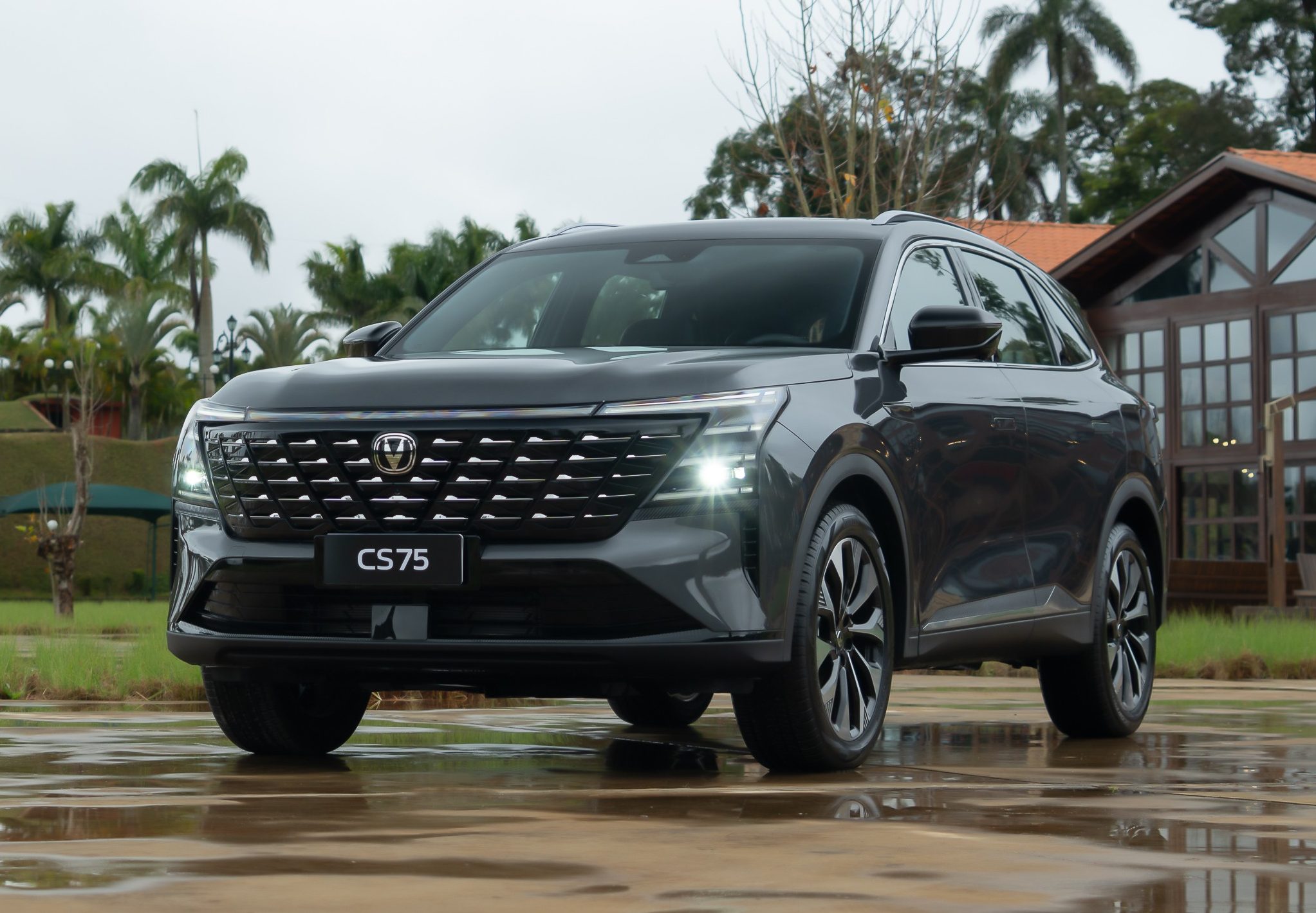 SUV Changan CS75 Plus cinza-escuro, com faróis acesos, estacionado em pátio molhado. Ao fundo, gramado, palmeiras, uma torre verde e uma casa de madeira com telhado de cerâmica
