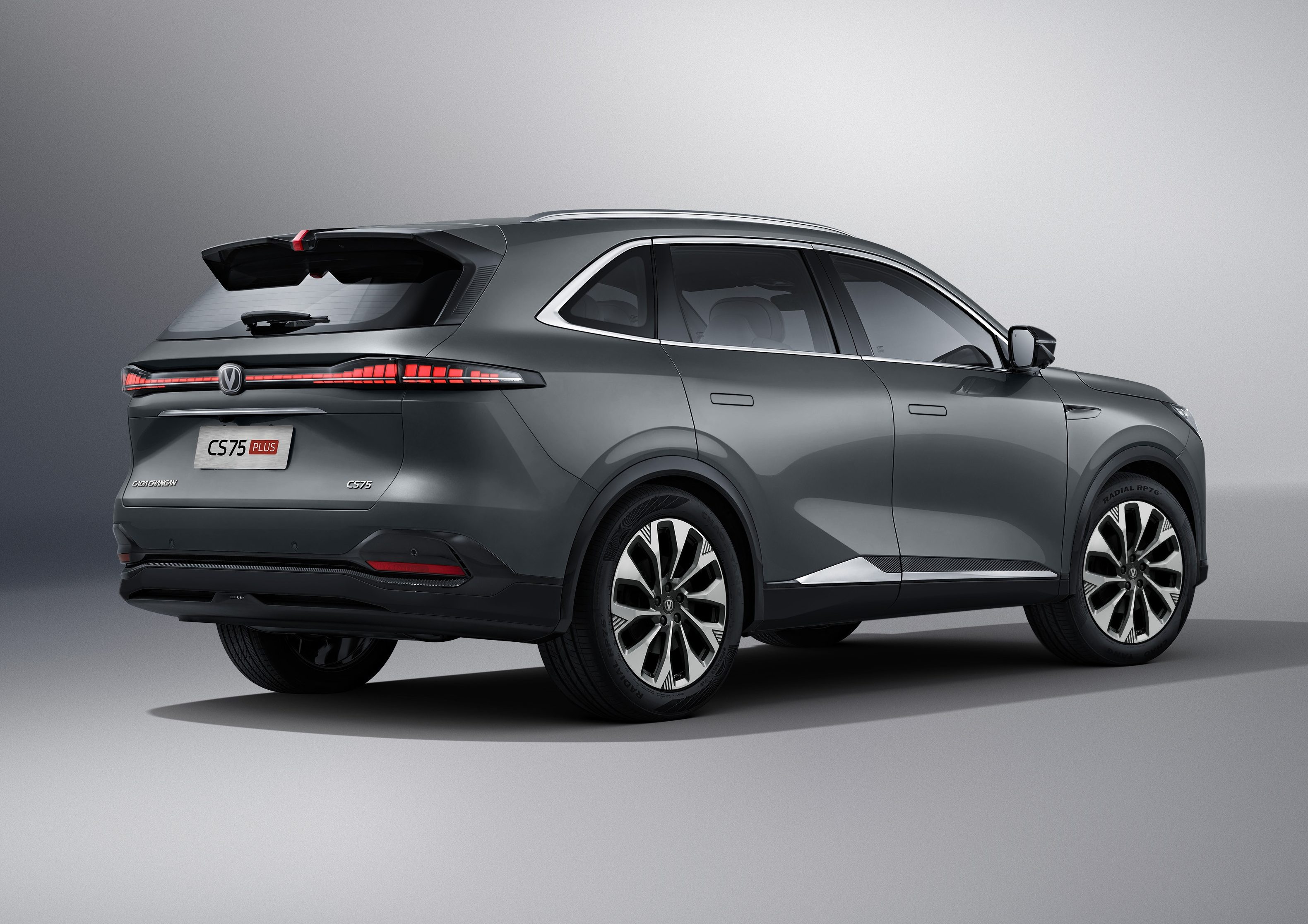 SUV cinza escuro, modelo Changan CS75 Plus, visto de traseira e lateral, parado em estúdio com fundo cinza claro. As lanternas traseiras estão acesas, e as rodas têm design de estrela em preto e prata