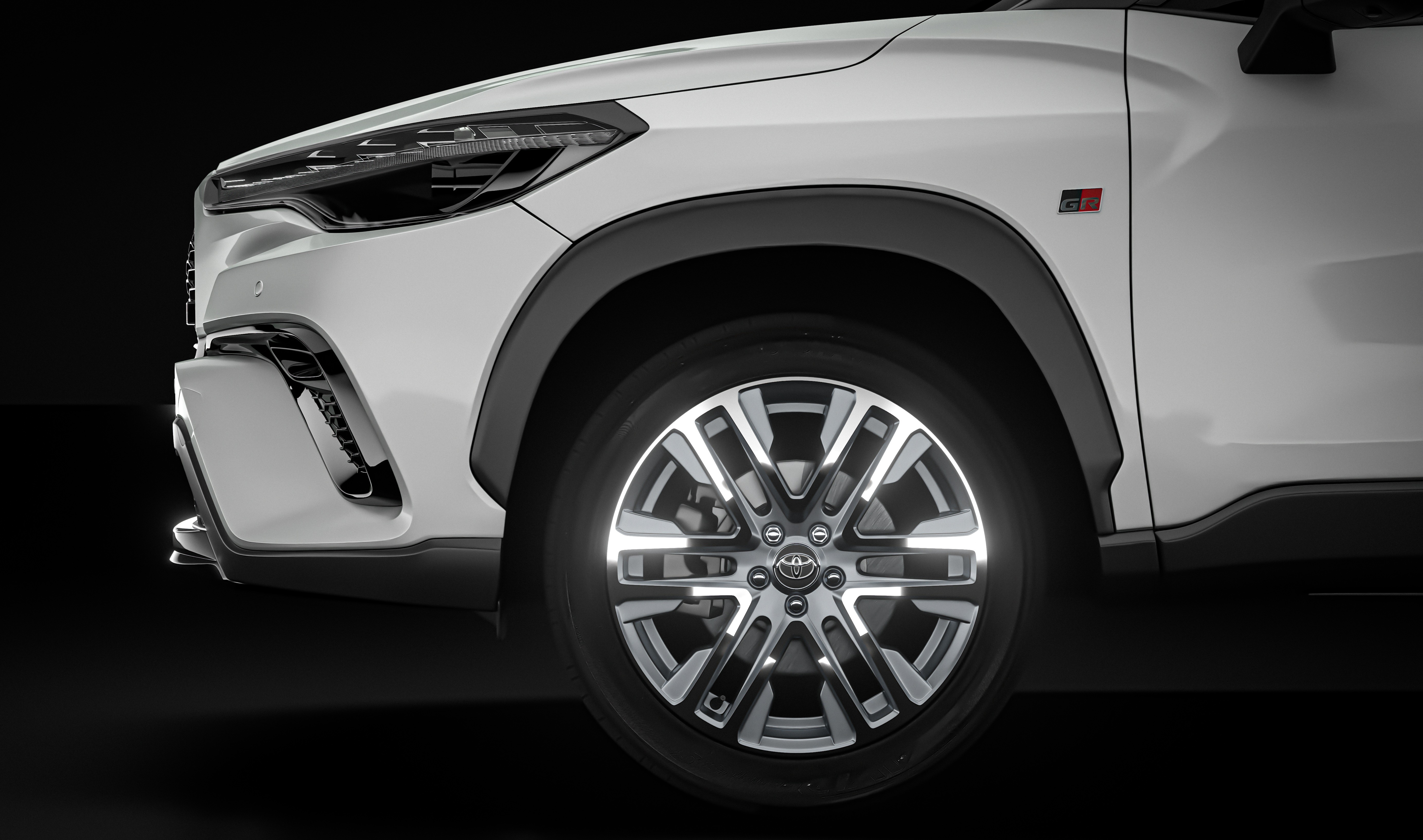 Lateral dianteira de um carro branco com detalhes pretos, incluindo o para-choque, a caixa de roda e a saia. A roda possui aros prateados e pretos, com o emblema da Toyota no centro. Um pequeno distintivo GR vermelho e preto está visível acima da roda, no para-lama. O fundo é totalmente preto