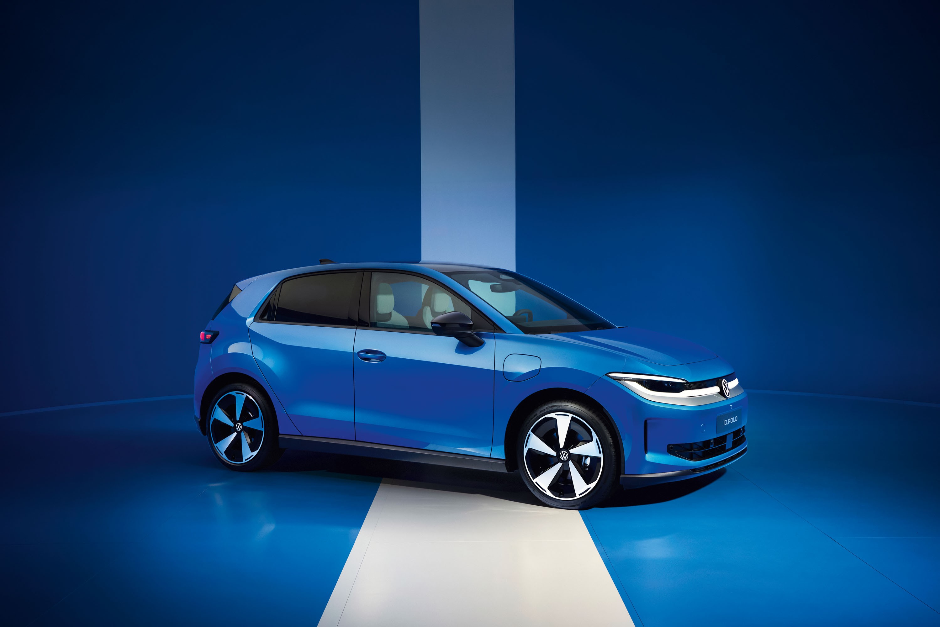 Carro elétrico azul brilhante, modelo hatchback, estacionado em um estúdio com paredes e chão azuis, e uma faixa branca vertical e horizontal cruzando o cenário. O veículo está levemente virado para a direita, exibindo suas rodas com design moderno em preto e branco. A iluminação destaca as linhas do carro e o ambiente monocromático