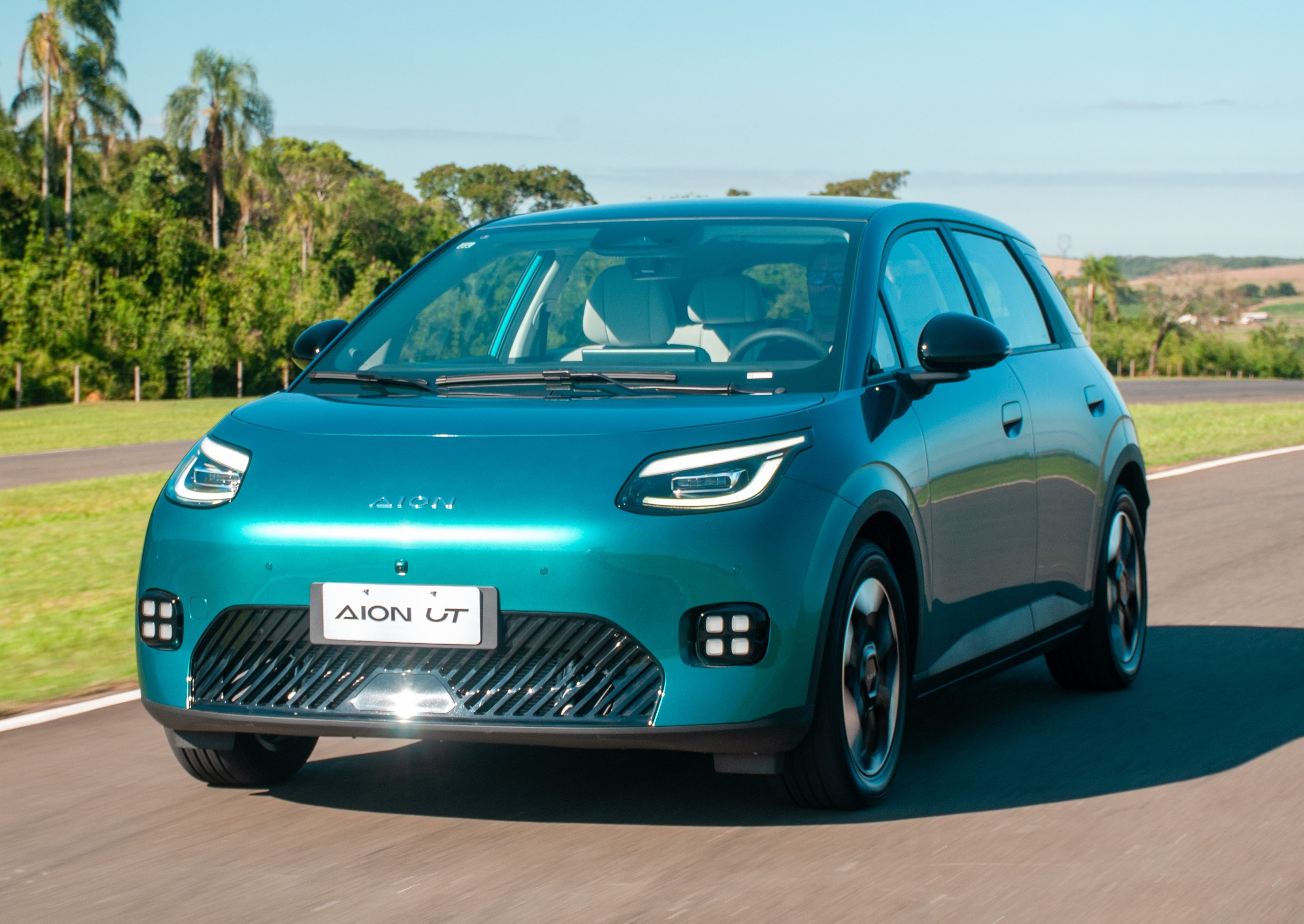 Carro elétrico Aion UT azul-turquesa em uma pista de asfalto, com vegetação exuberante e céu azul ao fundo