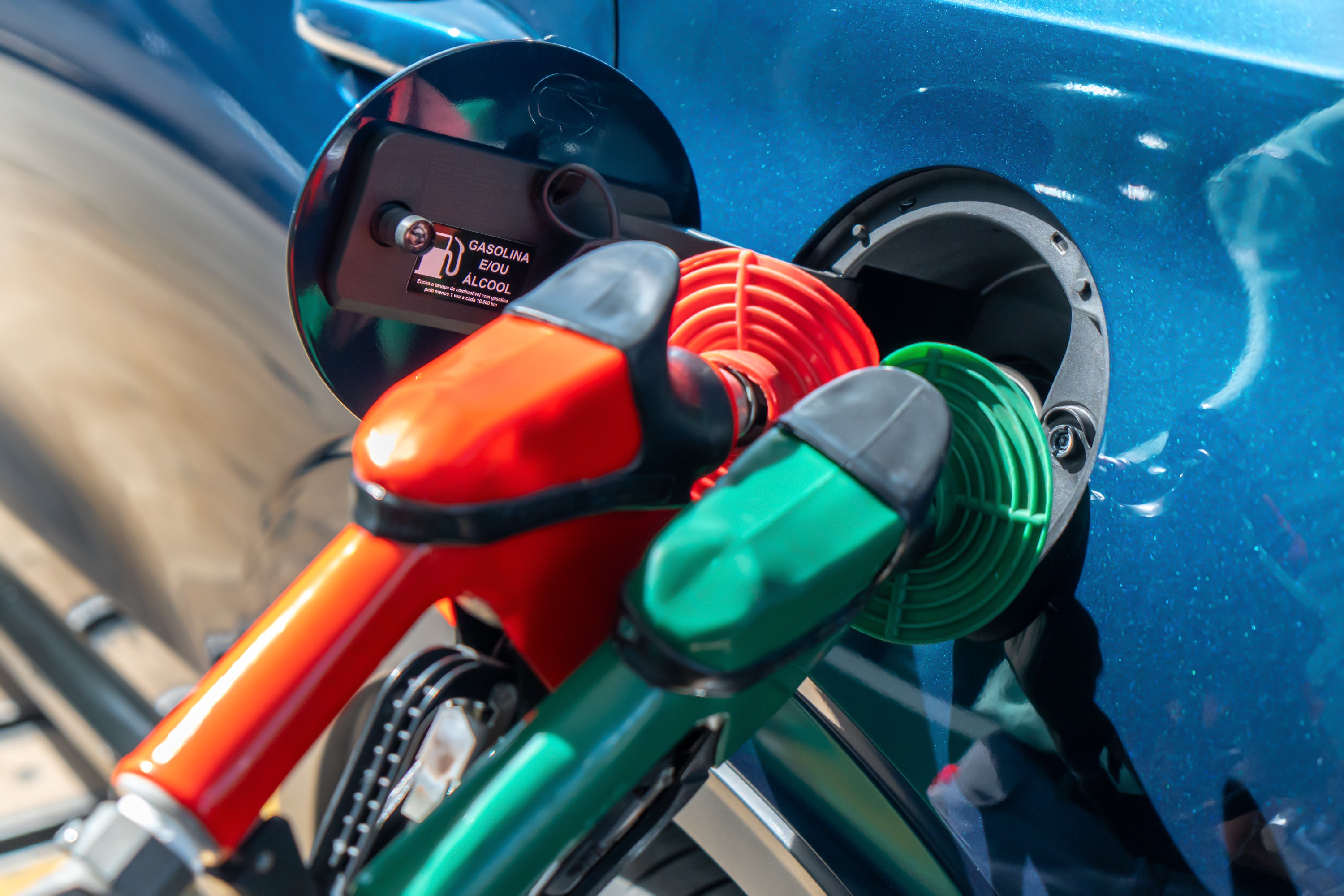 Bicos de bomba de combustível vermelho e verde inseridos no tanque de um carro azul, com a tampa aberta exibindo a inscrição GASOLINA E/OU ÁLCOOL