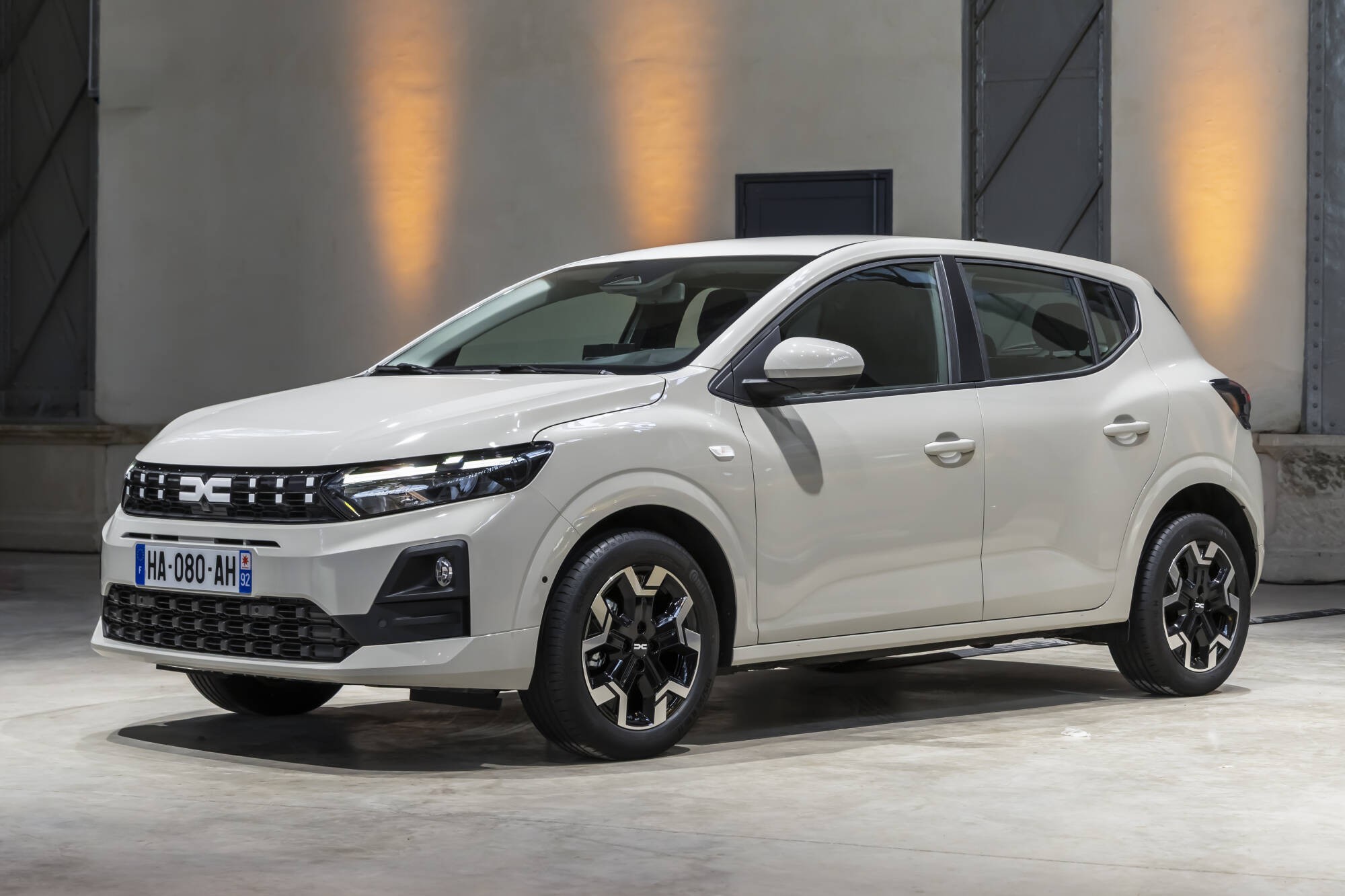 Carro branco Dacia Sandero Stepway, modelo 2024, estacionado em ambiente interno com parede cinza e iluminação amarela, visto de frente e lateral