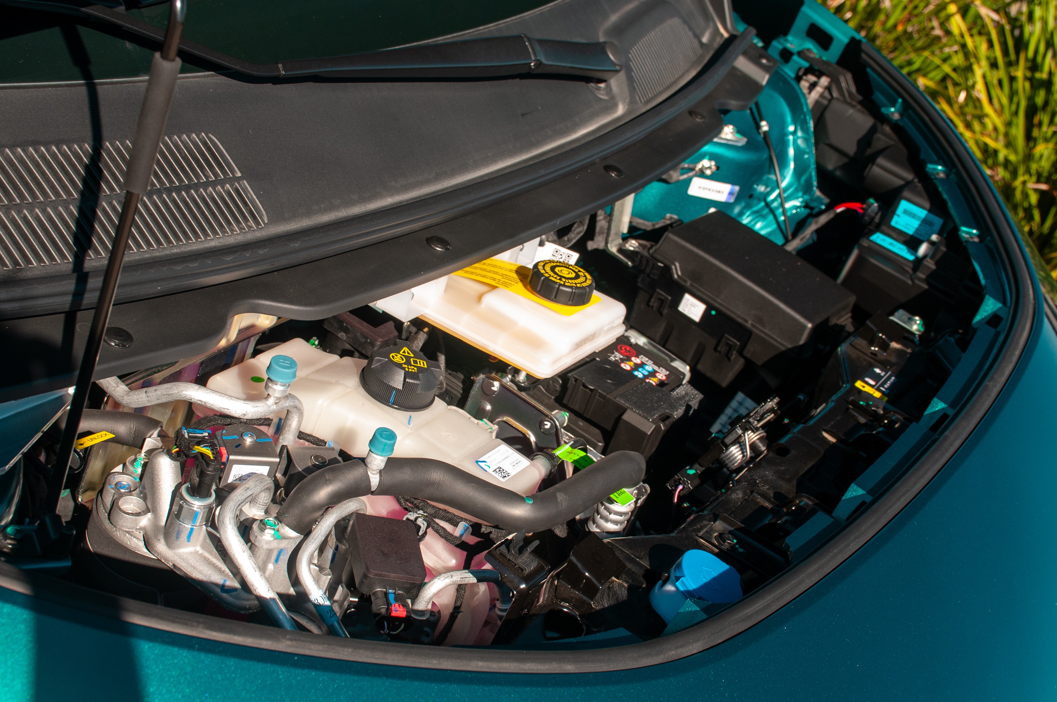 Compartimento do motor de um carro, com reservatórios de fluidos, cabos e caixas de fusíveis visíveis, em tons de preto, branco e amarelo, sobre a lataria verde-água do veículo