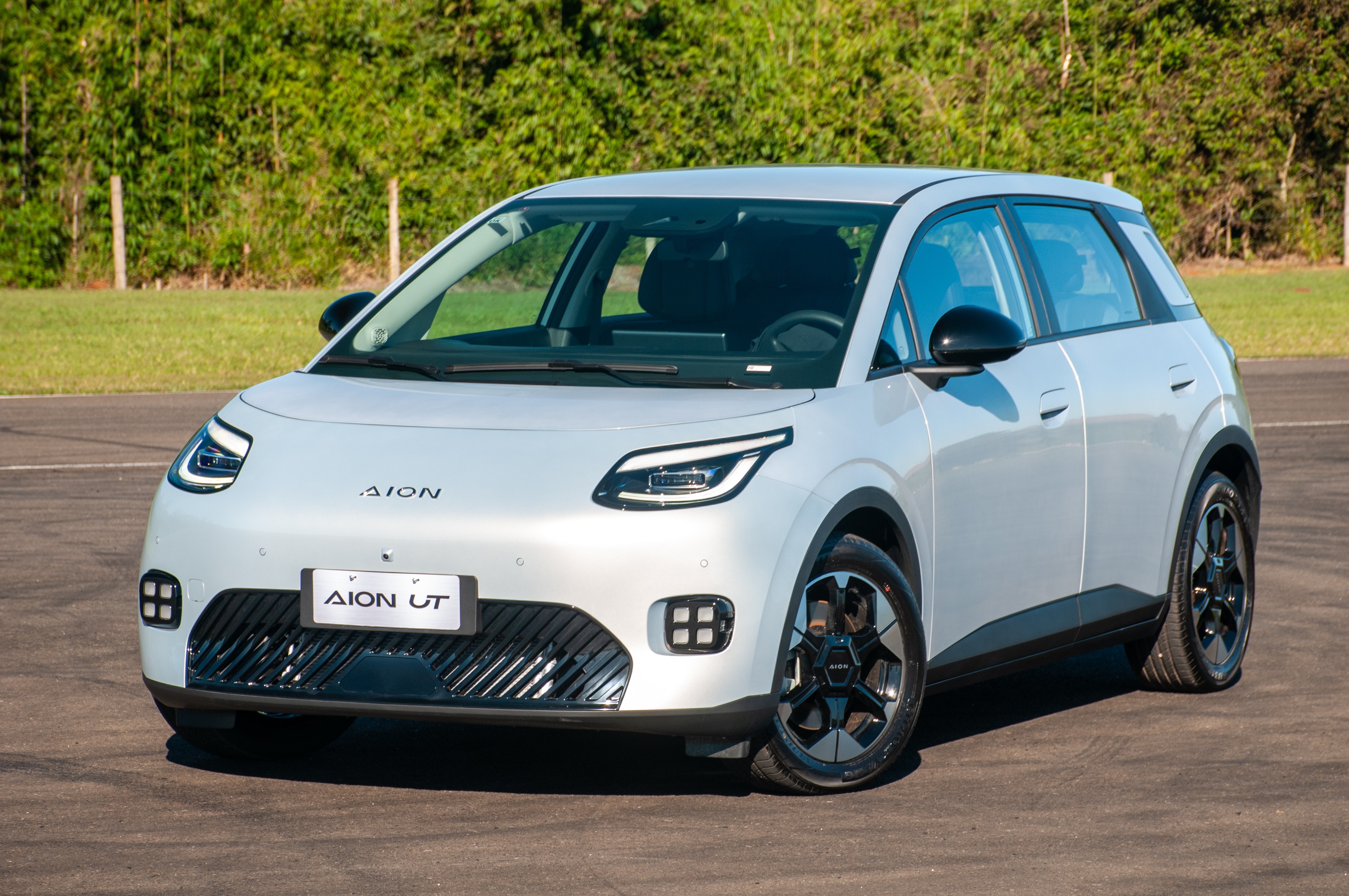 Carro elétrico Aion UT branco-prateado, com detalhes pretos nos retrovisores e rodas, estacionado em uma pista de asfalto com grama e árvores ao fundo