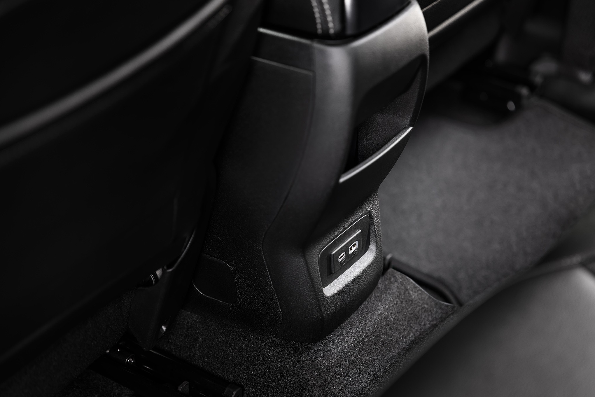 Detalhe do console central de um carro, com duas portas USB-A e USB-C para carregamento, localizadas na parte traseira, entre os bancos dianteiros. O interior é predominantemente preto, com estofamento e carpete escuros