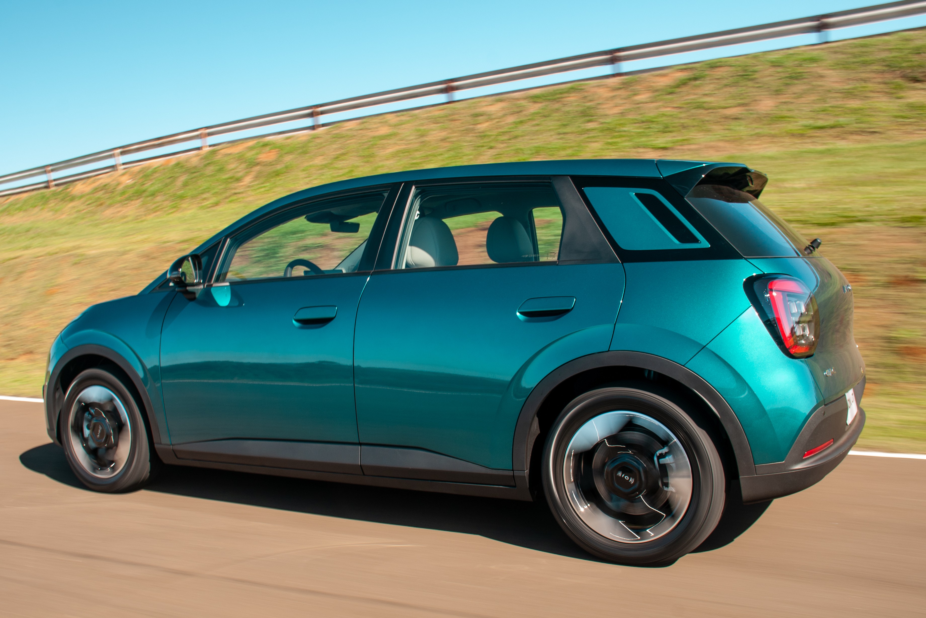 Carro SUV compacto azul-petróleo em movimento, visto de lado, com rodas pretas e detalhes em preto, em uma estrada com gramado e céu azul ao fundo