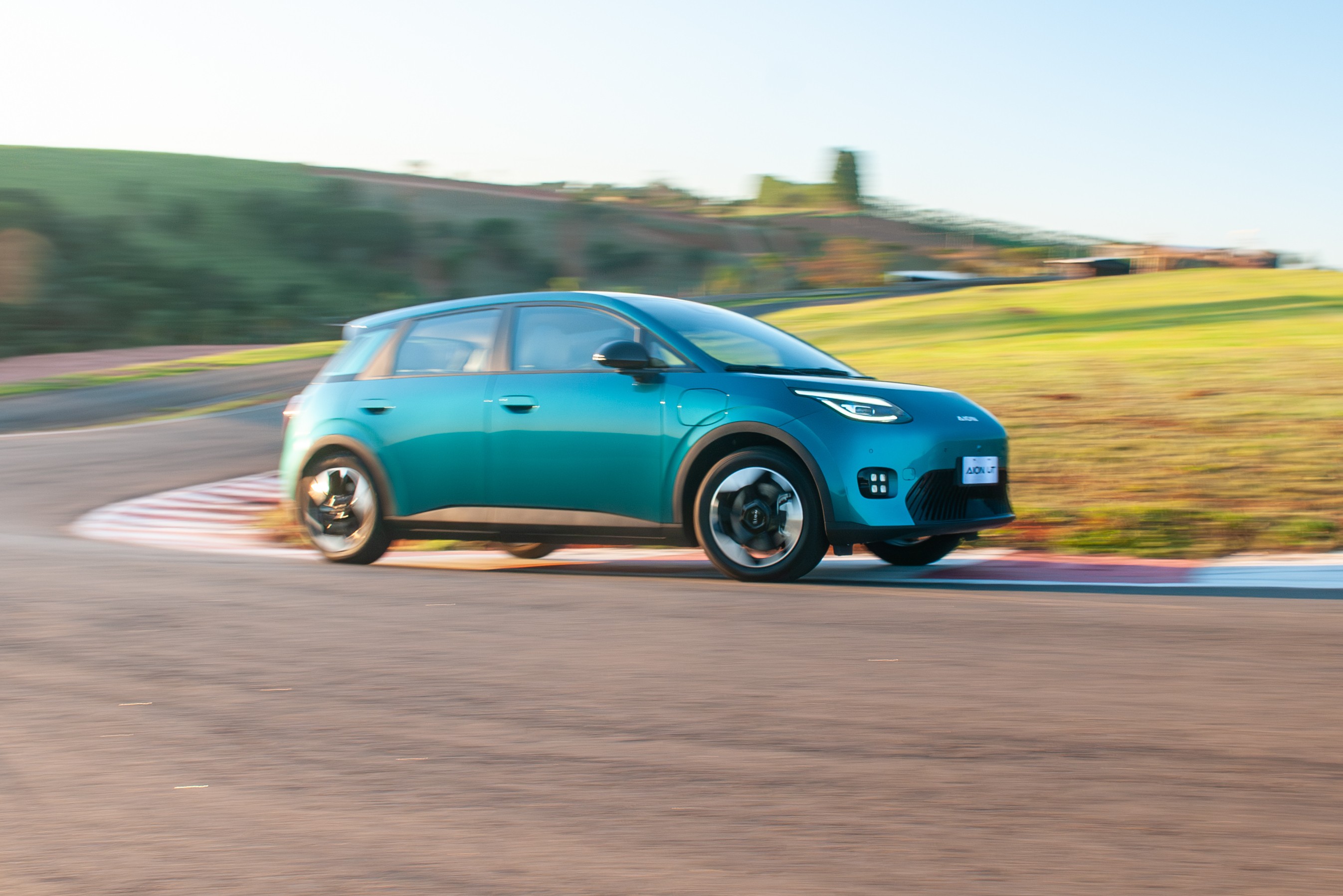 Carro elétrico azul-petróleo em movimento, fazendo uma curva acentuada em uma pista de corrida, com o fundo desfocado de colinas verdes e céu claro