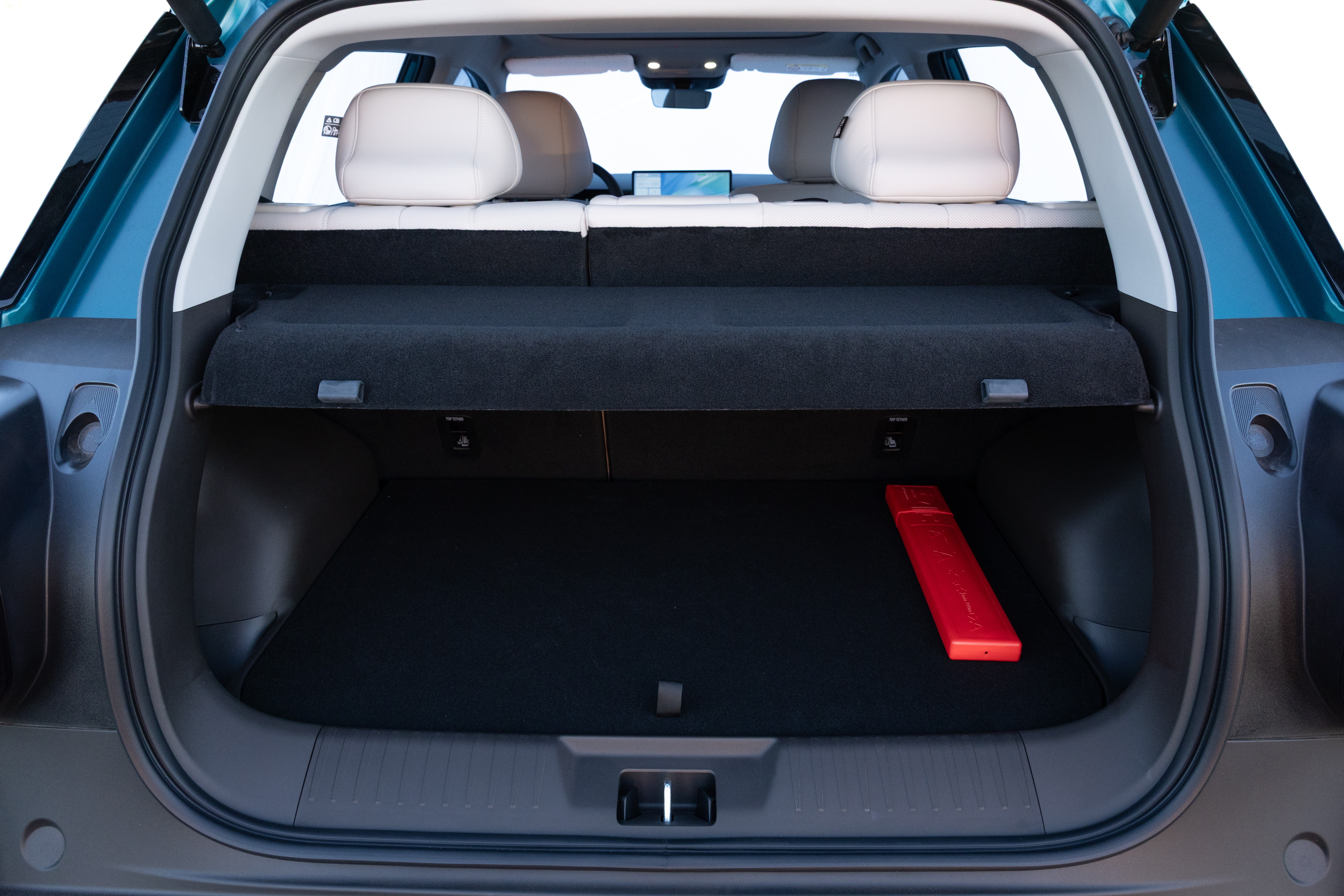 Porta-malas de um carro azul-petróleo, com o assoalho preto e um triângulo de sinalização vermelho no canto direito. O banco traseiro tem encostos de cabeça brancos e uma cobertura preta retrátil sobre o porta-malas