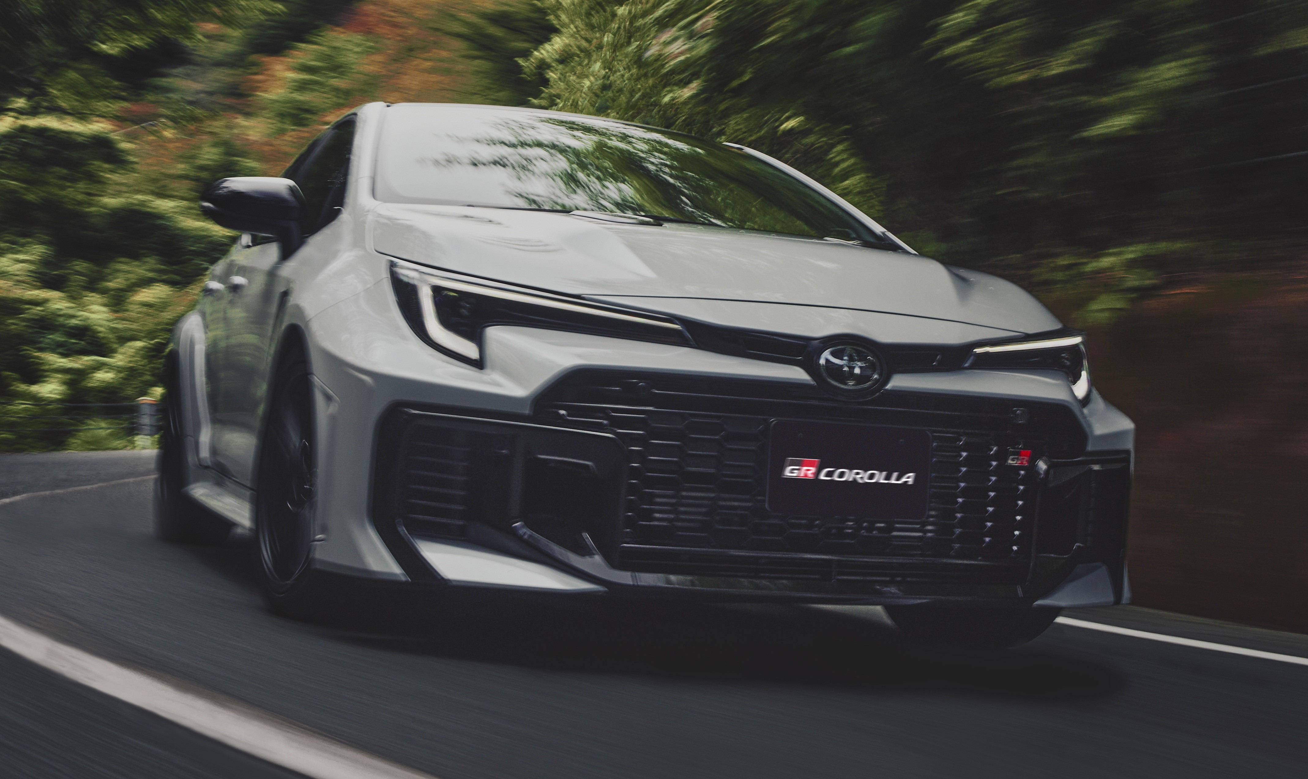 Carro Toyota GR Corolla cinza claro em movimento, visto de frente e levemente à direita, em uma estrada sinuosa com árvores borradas ao fundo