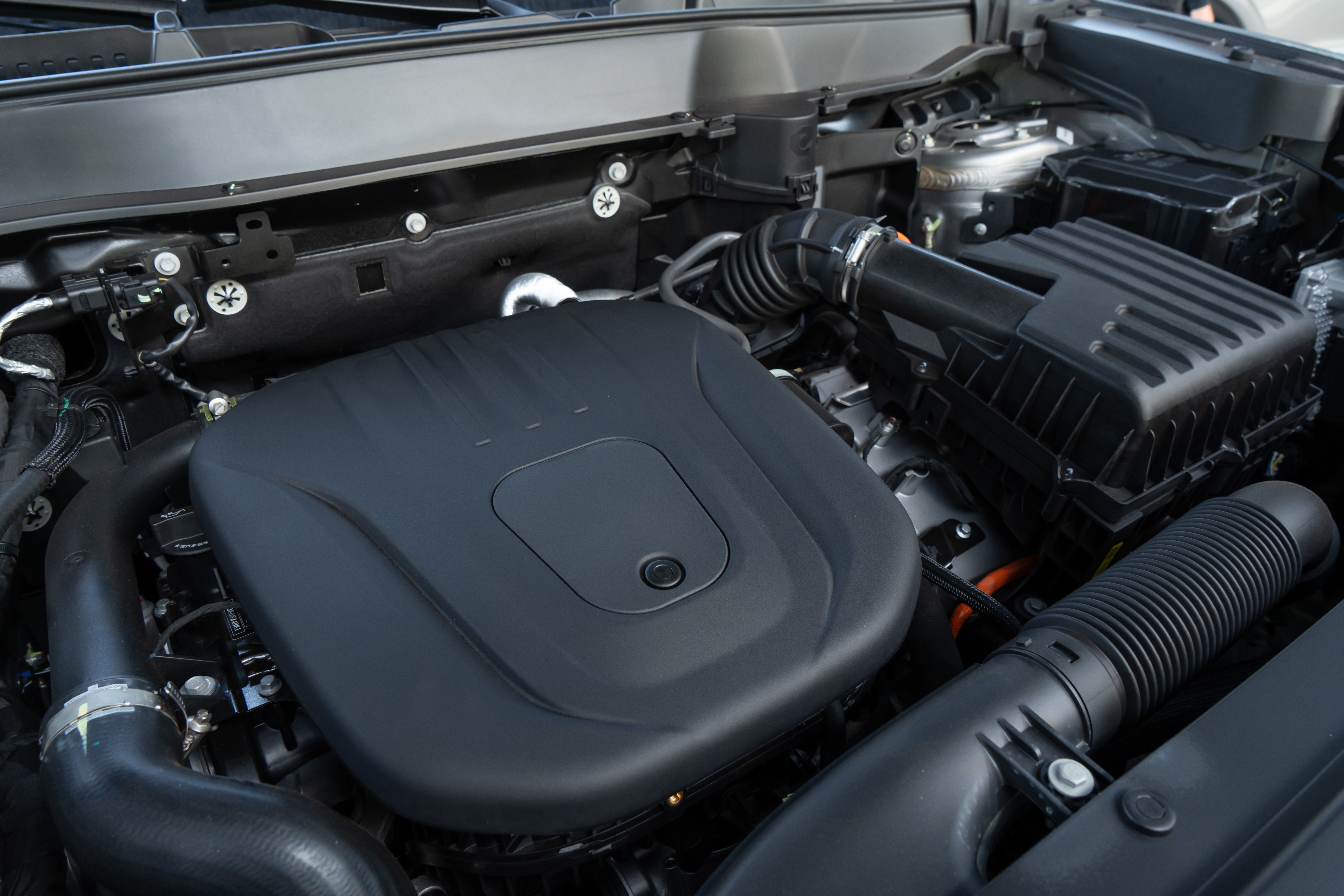 Vista superior do motor de um carro, com a tampa do motor preta e lisa no centro. Ao redor, tubulações, fios e componentes mecânicos em tons escuros, com detalhes prateados e alaranjados, indicando um veículo moderno