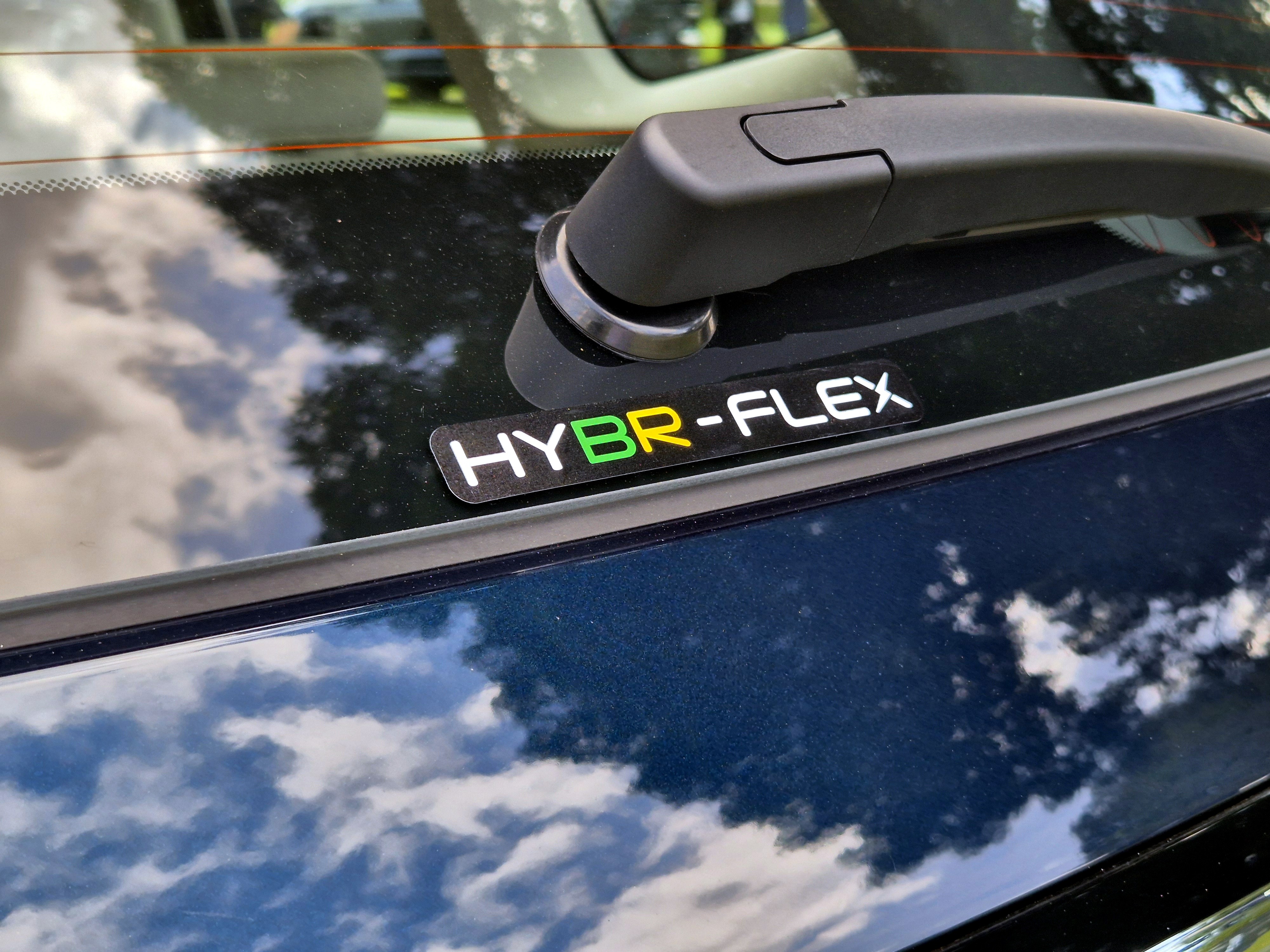 Adesivo HYBR-FLEX em preto com letras brancas, BR em verde e F em amarelo, colado no vidro traseiro de um carro azul escuro, abaixo do limpador de para-brisa. Nuvens e céu azul refletem na lataria e no vidro