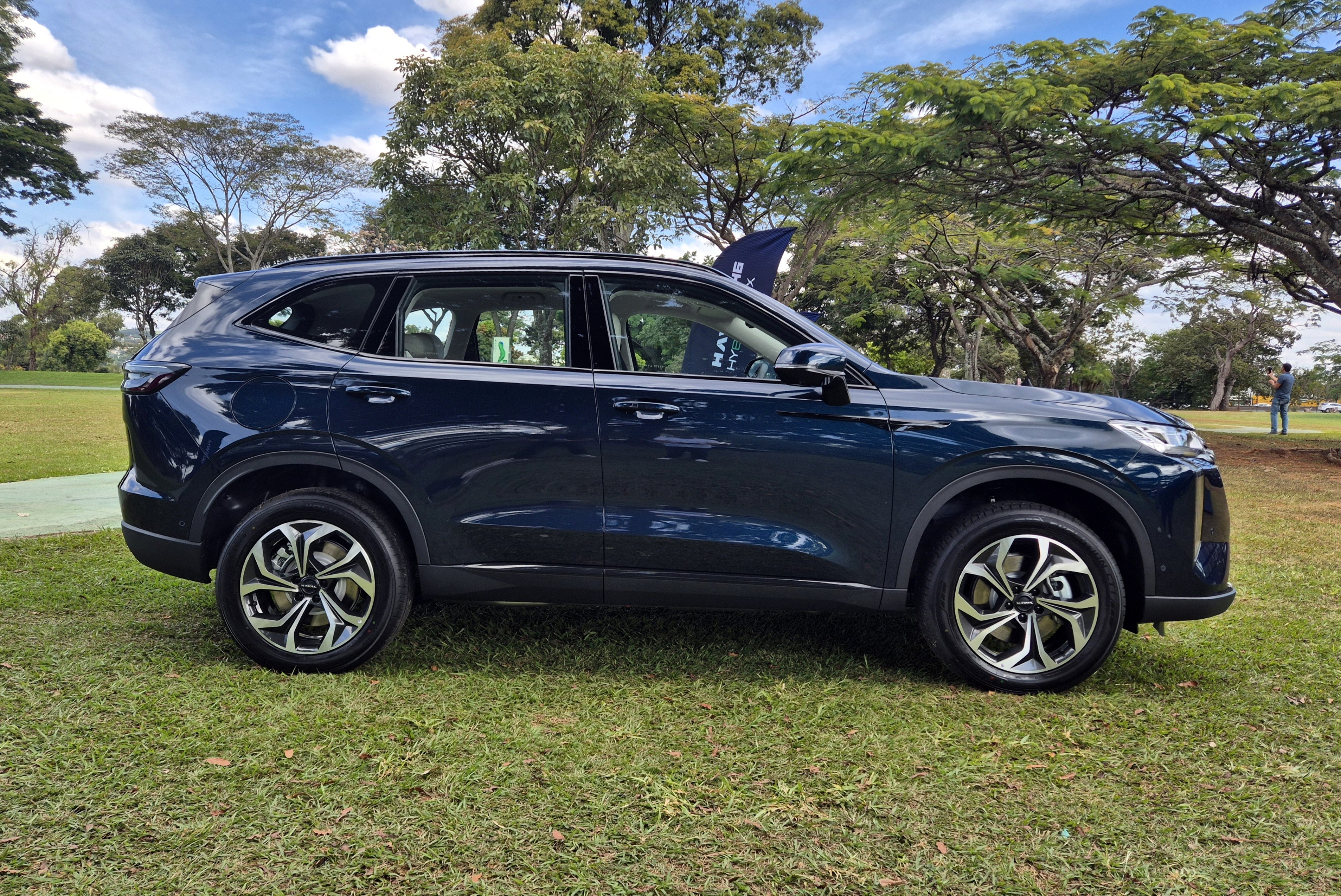 SUV azul-marinho estacionado na grama, com árvores e céu azul com nuvens ao fundo. Um homem distante aparece à direita