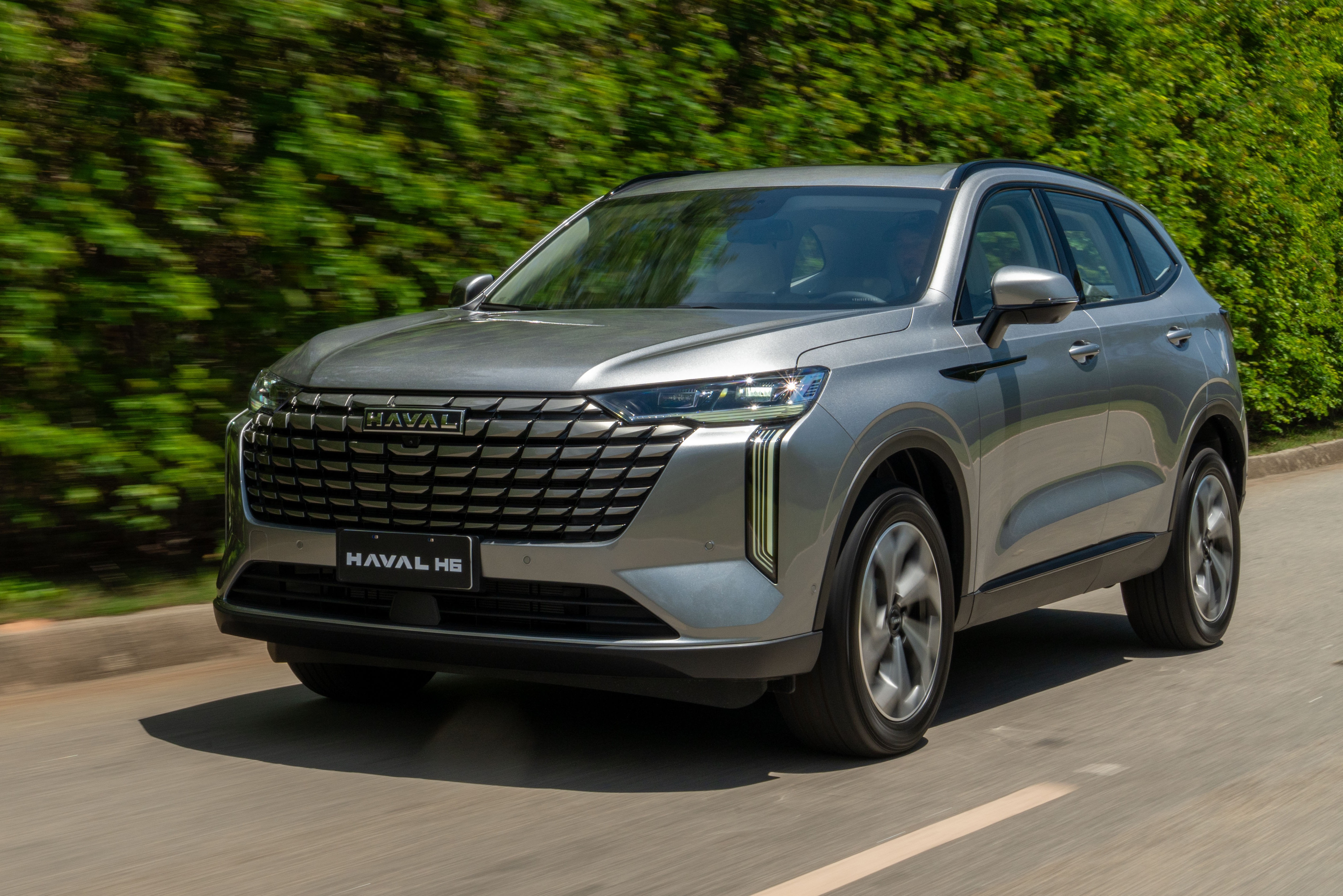 SUV Haval H6 cinza-prateado em movimento, visto de frente e levemente de lado, com grade frontal grande e faróis afilados. O carro está em uma estrada asfaltada com faixa branca, e há uma vegetação verde borrada ao fundo, indicando velocidade