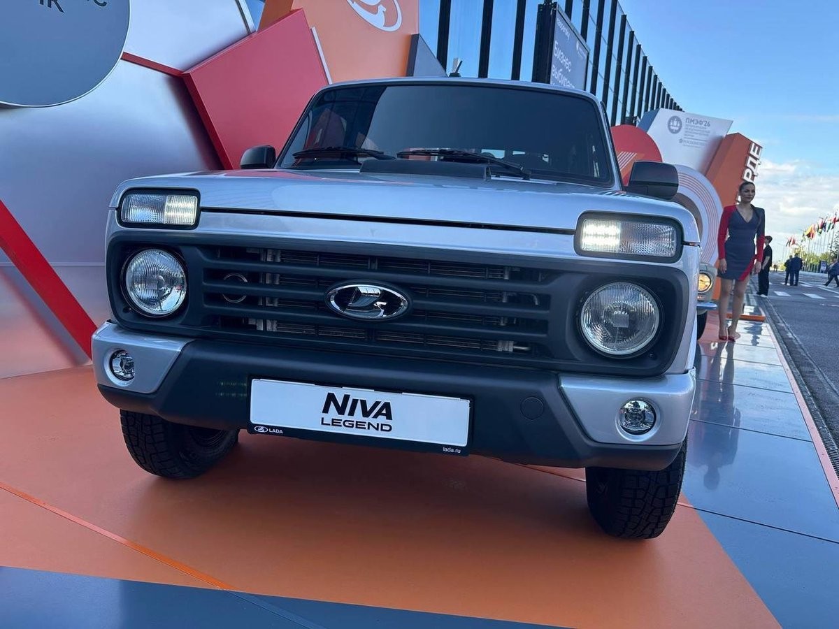 Carro Niva Legend prateado em exposição, visto de frente, com faróis acesos. Ao fundo, uma mulher de vestido vermelho e preto, e estruturas geométricas coloridas