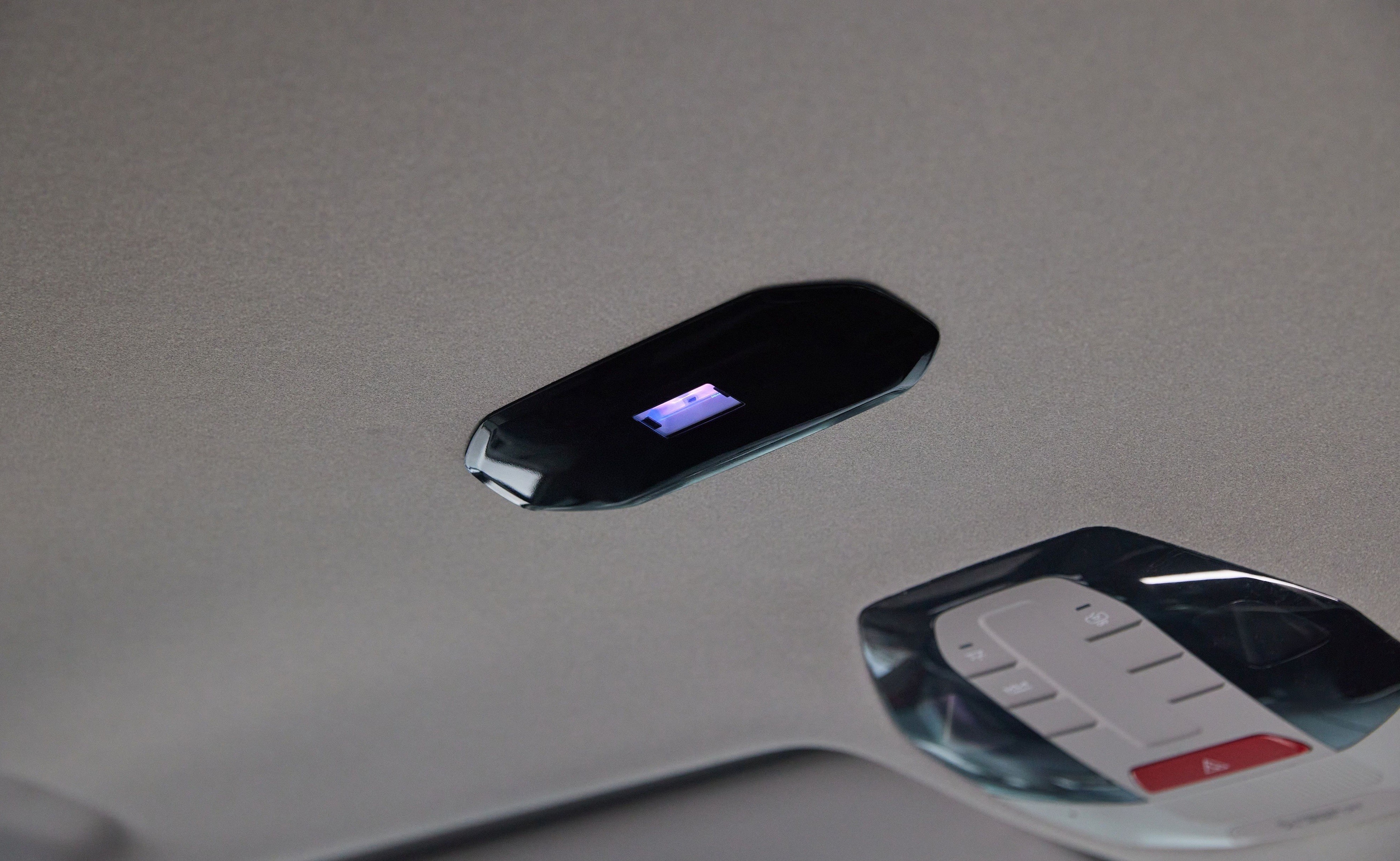 Painel de controle no teto de um carro, com uma tela retangular preta exibindo luz azul e um conjunto de botões brancos e um vermelho