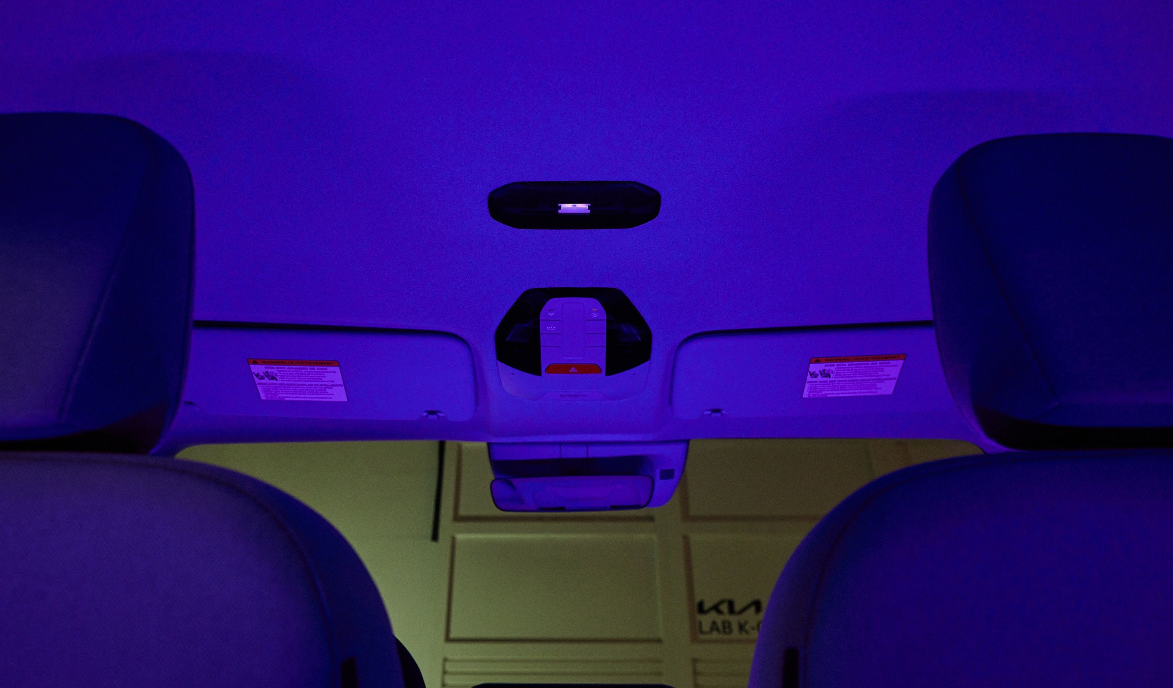 Interior de um carro com iluminação ambiente roxa, vista de trás dos bancos dianteiros. O teto e os para-sóis estão visíveis, com uma luz de teto central e um espelho retrovisor. Ao fundo, uma parede clara com o logo KIA LAB K aparece
