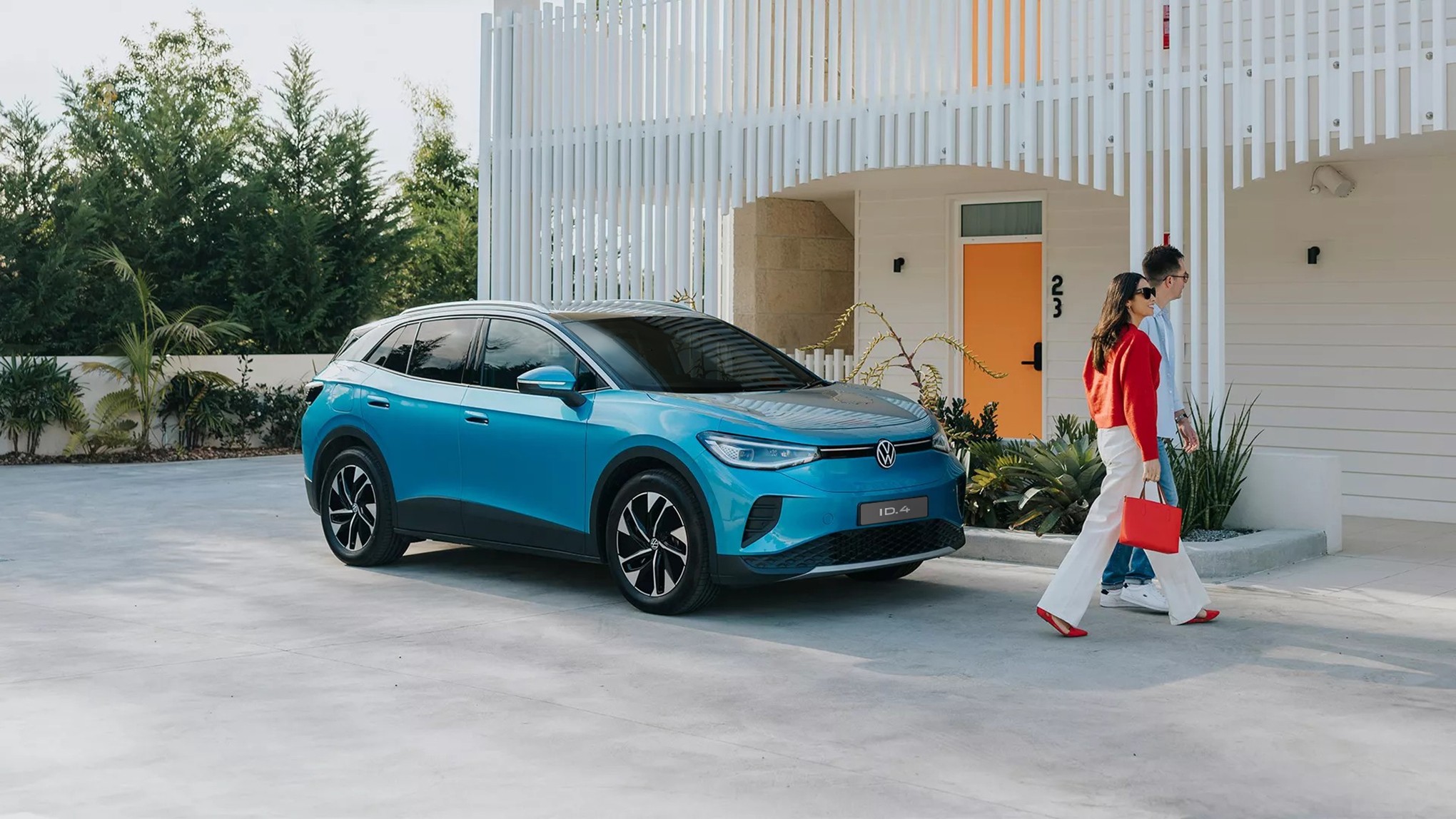 Carro elétrico Volkswagen ID.4 azul estacionado em frente a um prédio branco com ripas verticais. Um casal caminha à direita, a mulher de vermelho e calça branca, o homem de camisa clara e jeans