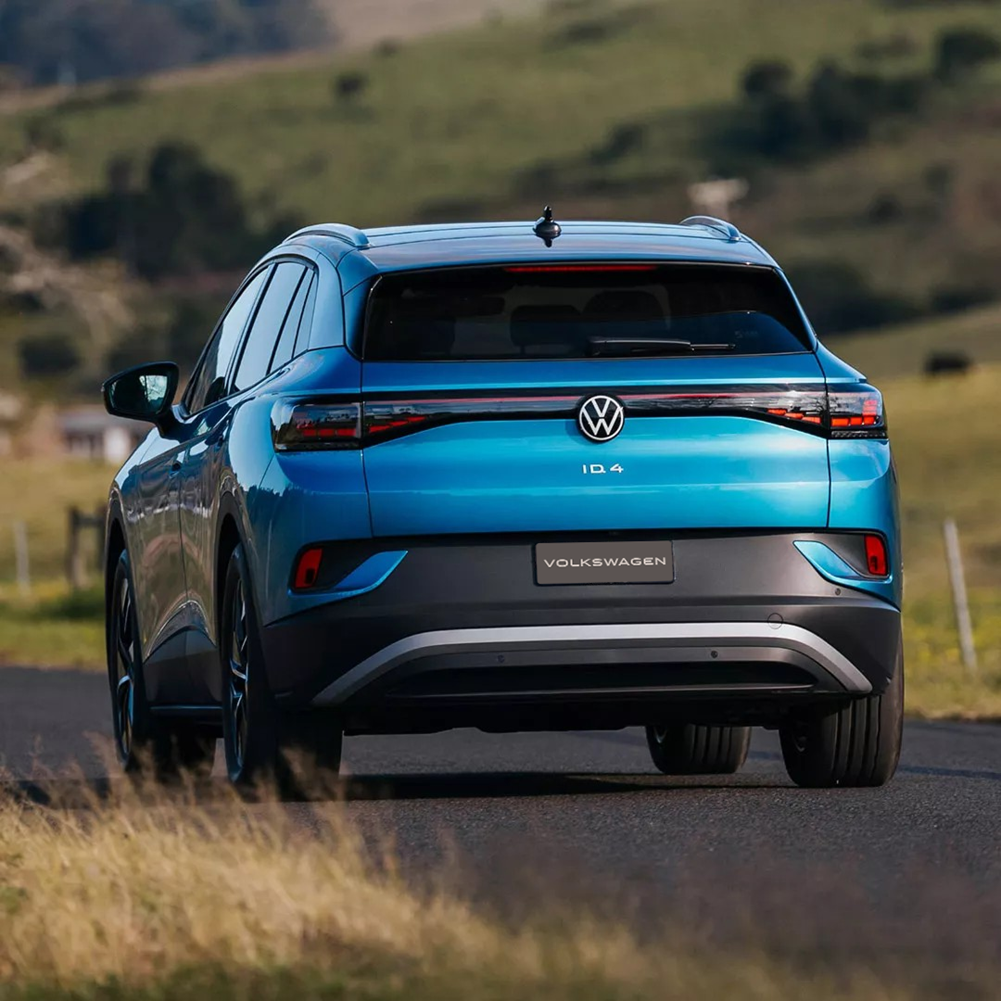 Traseira de um carro SUV elétrico Volkswagen ID.4 azul, visto de levemente à direita, em uma estrada asfaltada. Ao fundo, colinas verdes e céu claro. O carro tem lanternas traseiras conectadas por uma barra preta e o logotipo da Volkswagen no centro