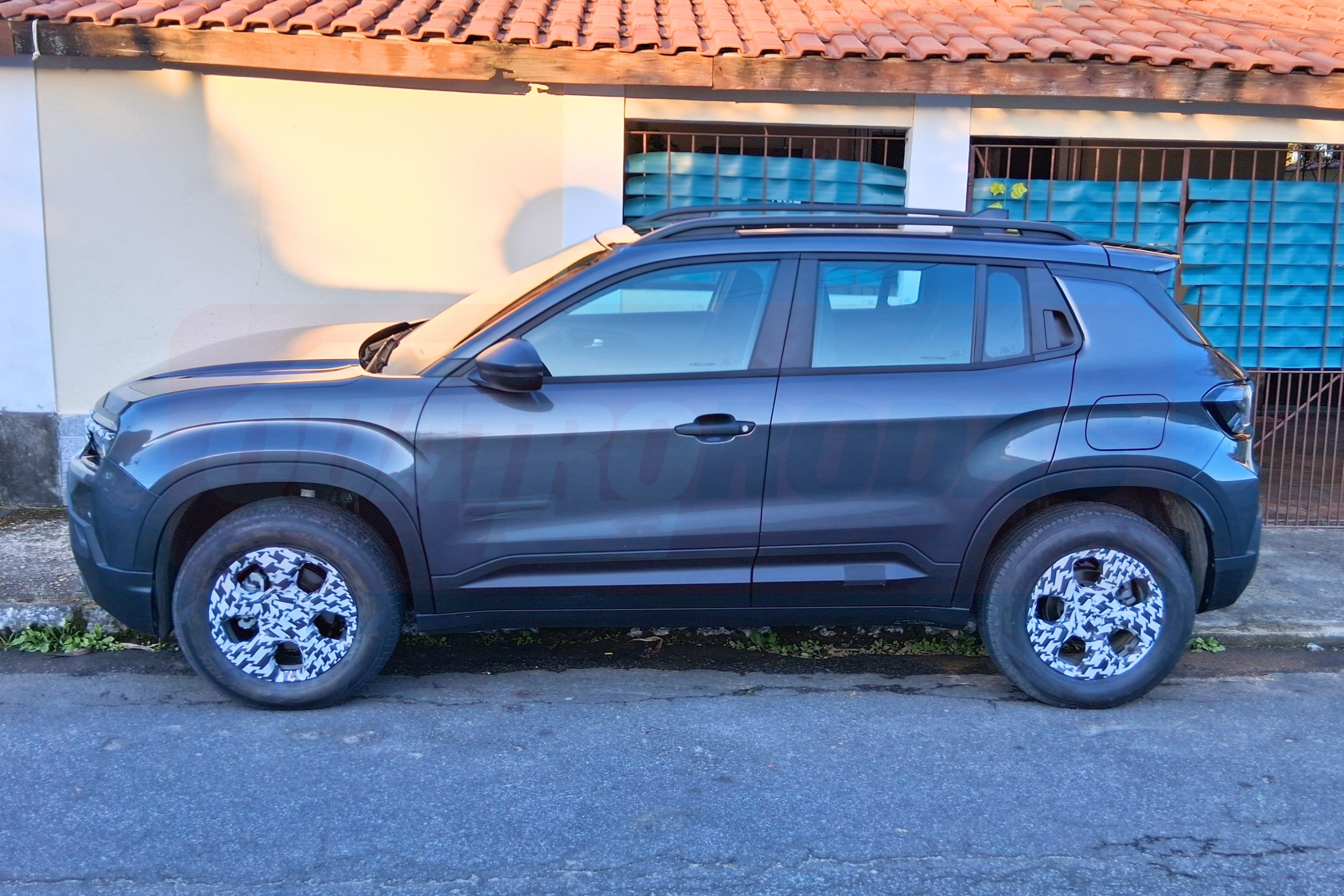 SUV cinza escuro estacionado na rua, com calotas camufladas em preto e branco, em frente a uma casa com telhado de cerâmica e portão azul
