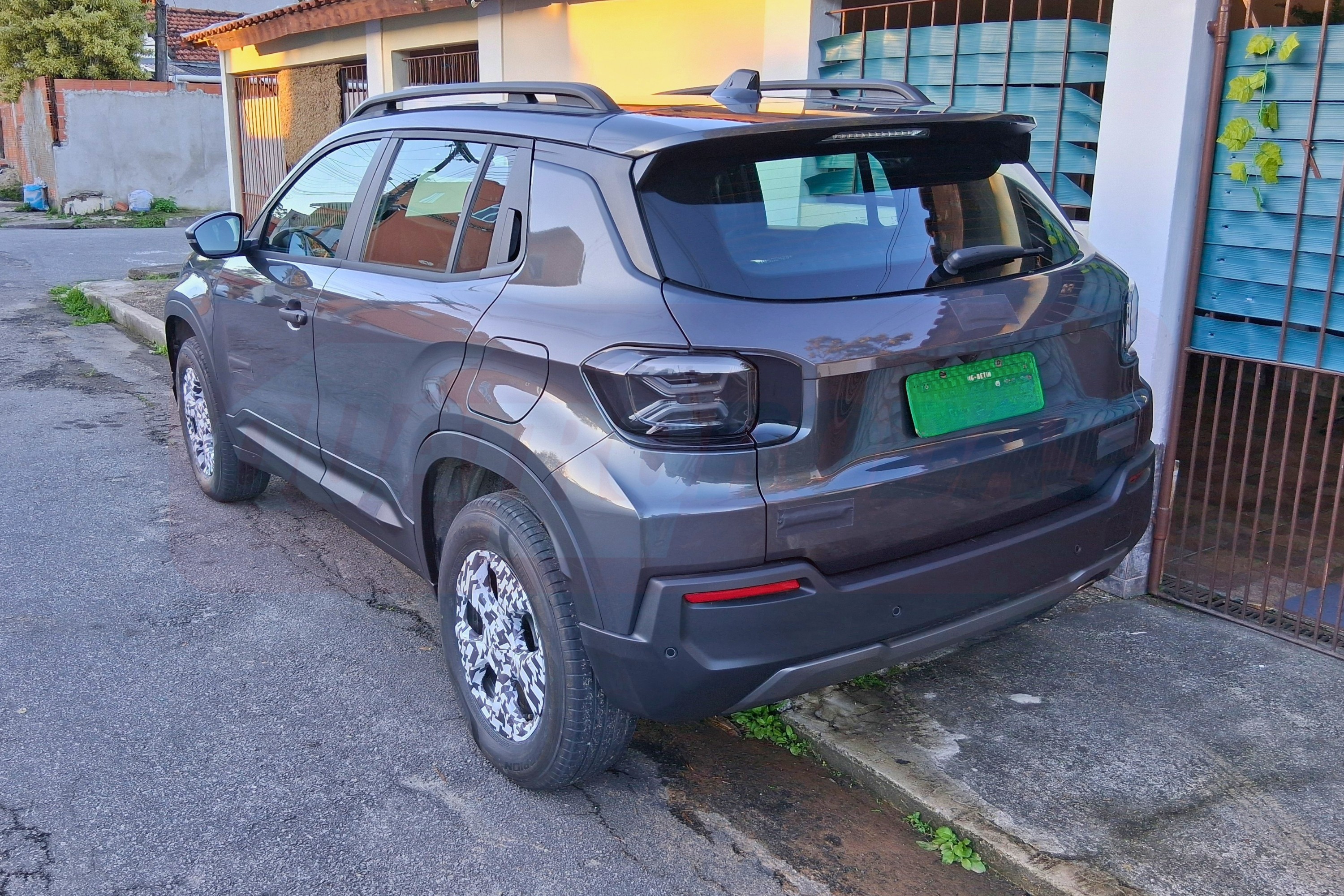 SUV cinza escuro estacionado na rua, com teto solar e rack de teto, visto de trás para a direita. A placa verde indica que é um veículo elétrico. Ao fundo, casas e um portão azul.