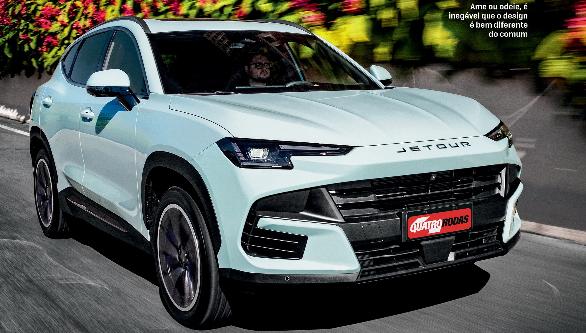 SUV Jetour branco-azulado em movimento na rua, com um homem dirigindo, e vegetação colorida ao fundo. O carro tem o nome JETOUR no capô e um emblema QUATRO RODAS na grade frontal. O texto superior direito diz: Ame ou odeie, é inegável que o design é bem diferente do comum