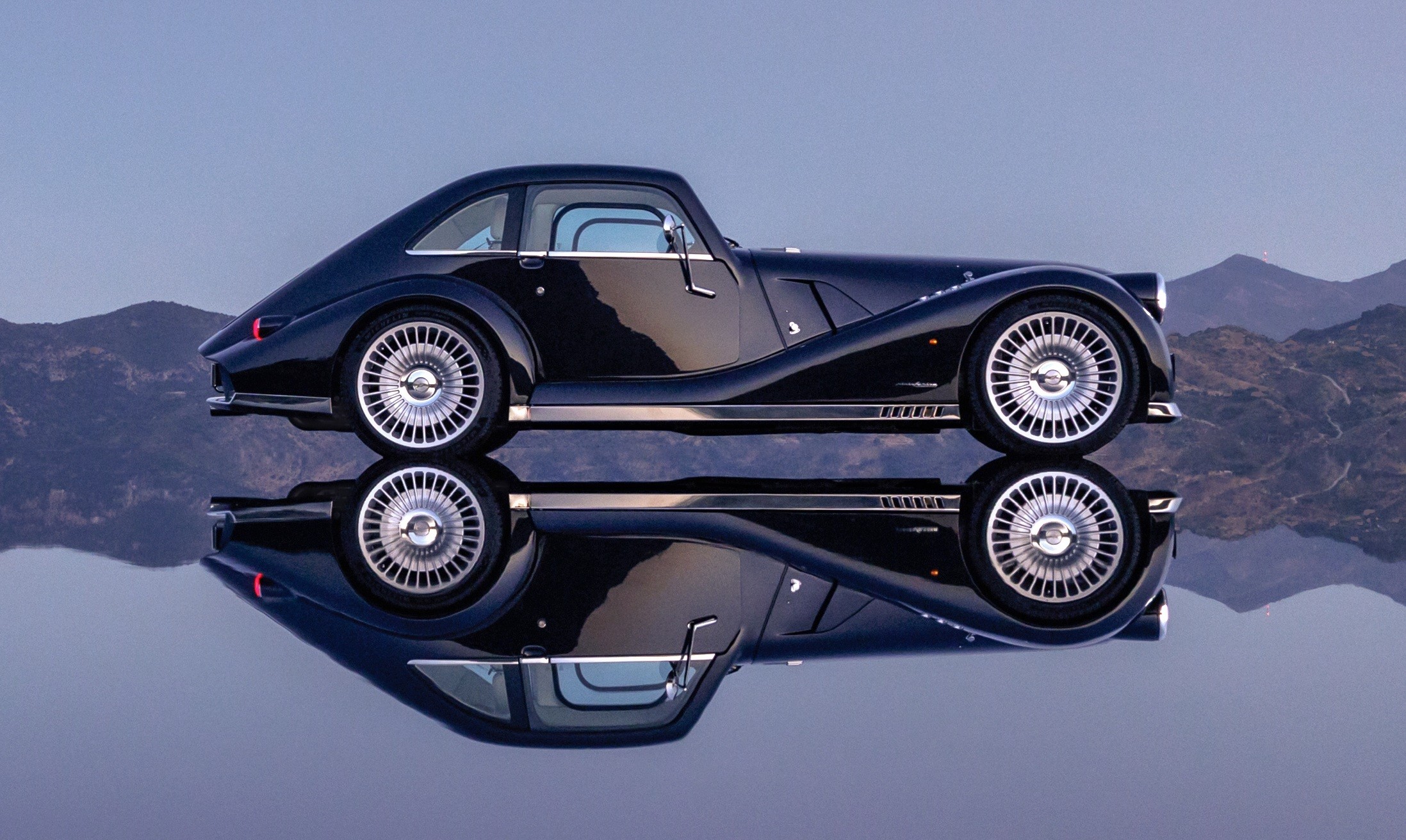 Carro esportivo preto vintage com rodas prateadas, refletido em uma superfície espelhada, com montanhas ao fundo sob céu azul claro