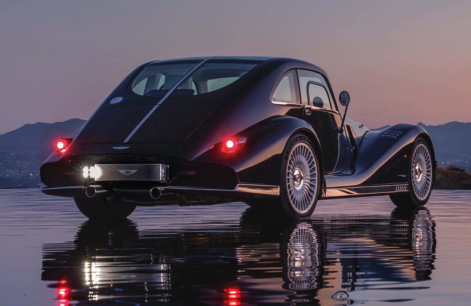 Carro esportivo Morgan Plus Four preto, com design retrô, visto por trás e ligeiramente de lado, parado em uma superfície molhada que reflete suas luzes traseiras vermelhas e a placa iluminada. Ao fundo, montanhas escuras sob um céu crepuscular em tons de roxo e laranja
