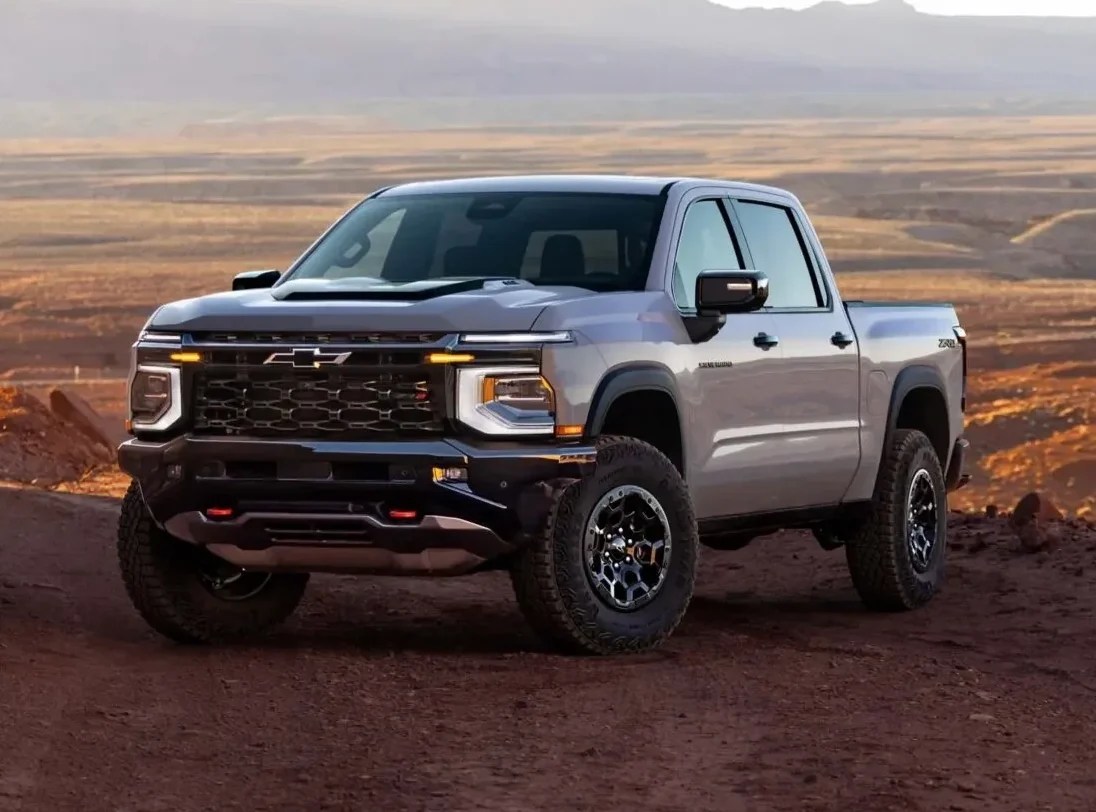 Picape Chevrolet Silverado cinza clara em uma estrada de terra avermelhada, com montanhas e um vasto deserto ao fundo sob um céu claro