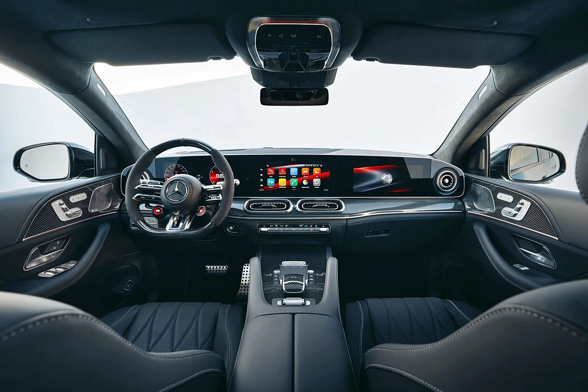 Interior de um carro Mercedes-Benz, com o volante esportivo em camurça e detalhes vermelhos. O painel digital exibe informações do veículo e um sistema multimídia com ícones coloridos. Os bancos de couro preto têm costura branca e o console central possui acabamento em fibra de carbono. O teto solar e os espelhos retrovisores são visíveis