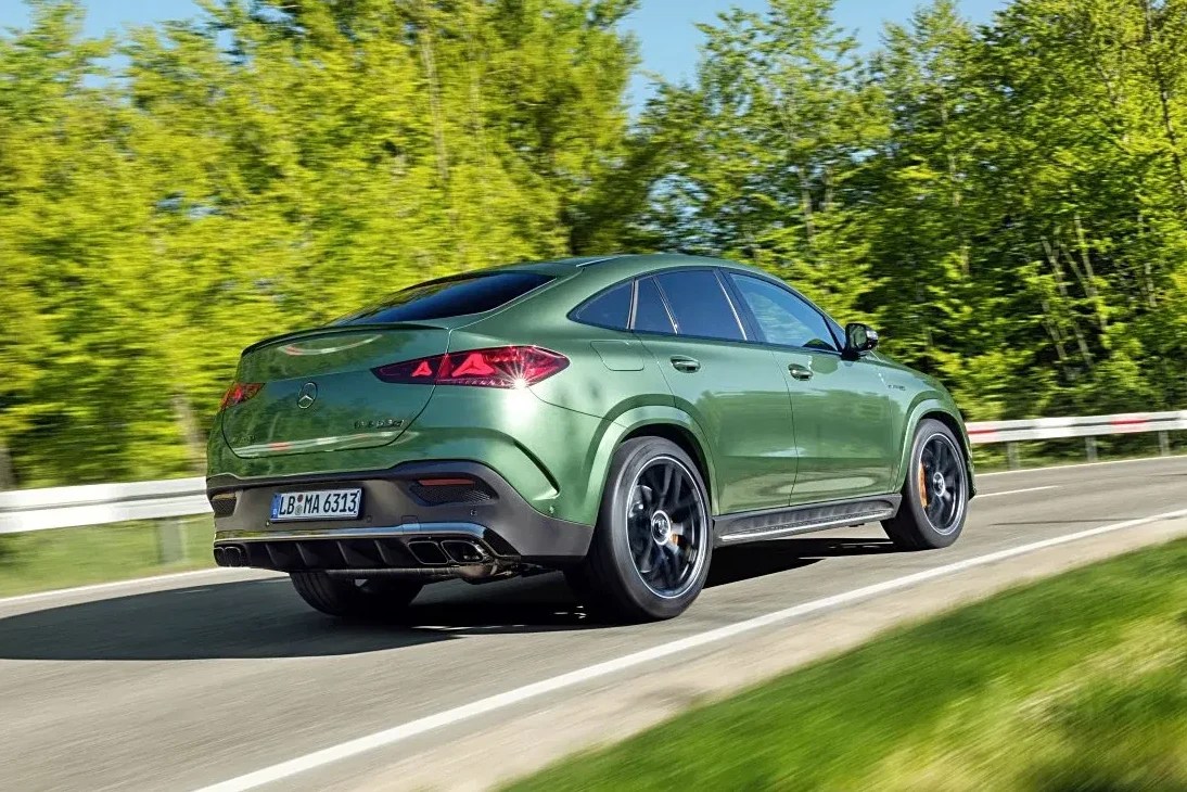 SUV cupê verde escuro em movimento, visto por trás, em uma estrada asfaltada com faixa branca. À direita, gramado e guarda-corpo metálico. Ao fundo, árvores com folhagem verde vibrante sob céu azul claro