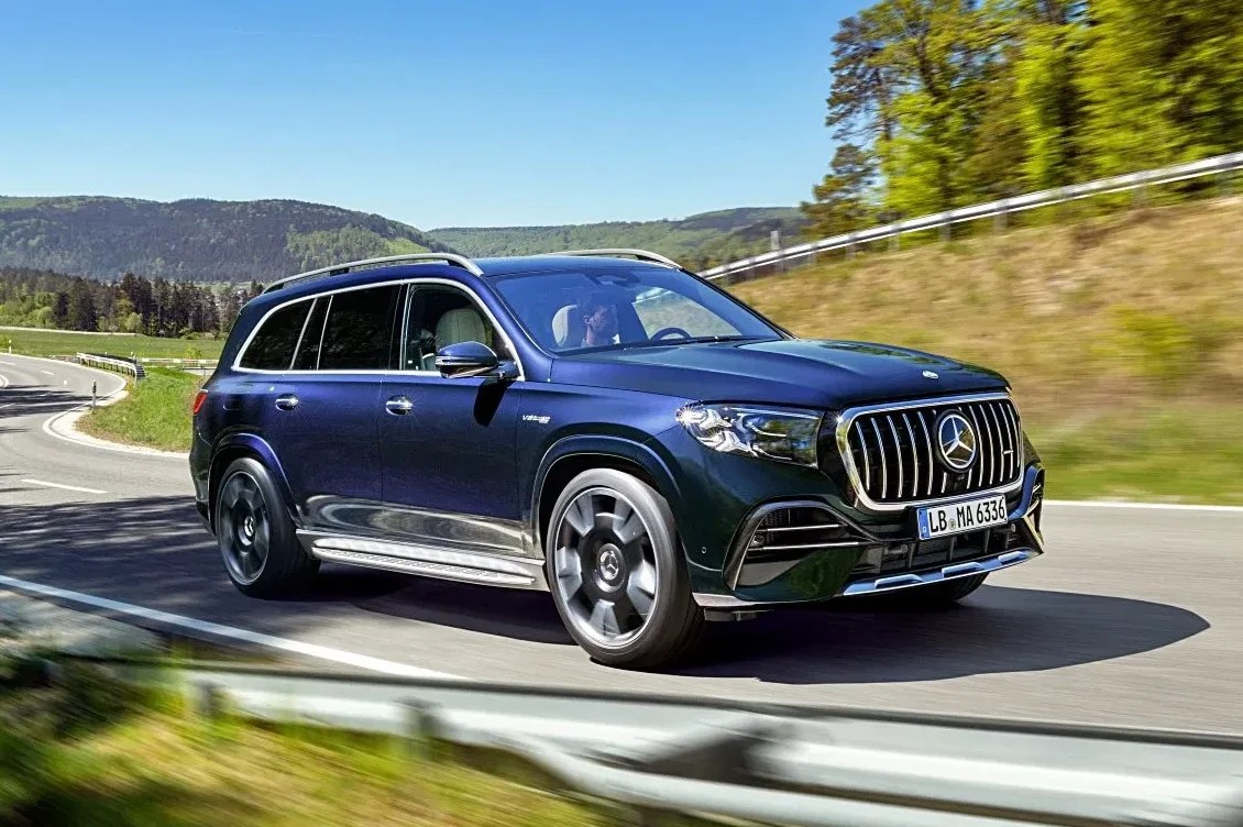 SUV Mercedes-Benz GLS azul escuro em movimento numa estrada sinuosa, com árvores verdes e montanhas ao fundo sob um céu azul claro