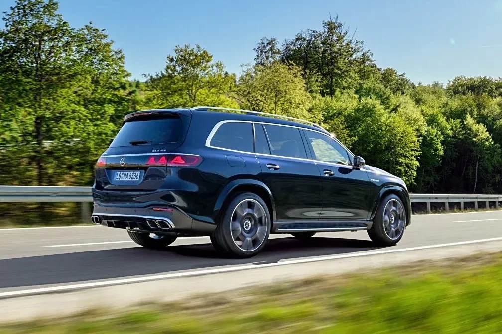 SUV Mercedes-Benz GLS 63 AMG preto em movimento numa estrada sinuosa, com árvores e gramados verdes ao redor, sob céu azul claro e sol forte