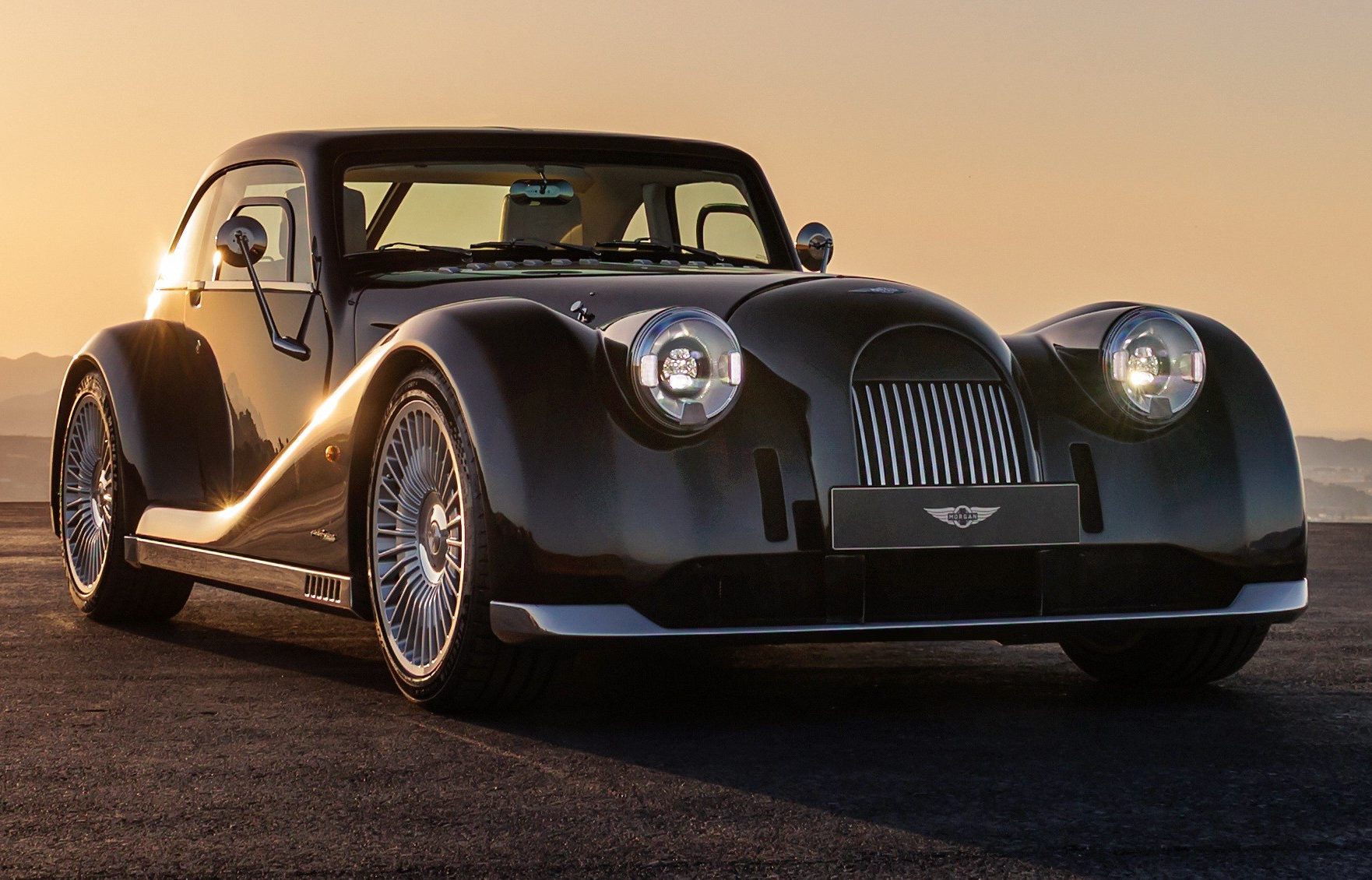 Carro esportivo Morgan Plus Four preto brilhante, com faróis acesos e rodas prateadas, estacionado em uma estrada escura ao pôr do sol, com montanhas ao fundo