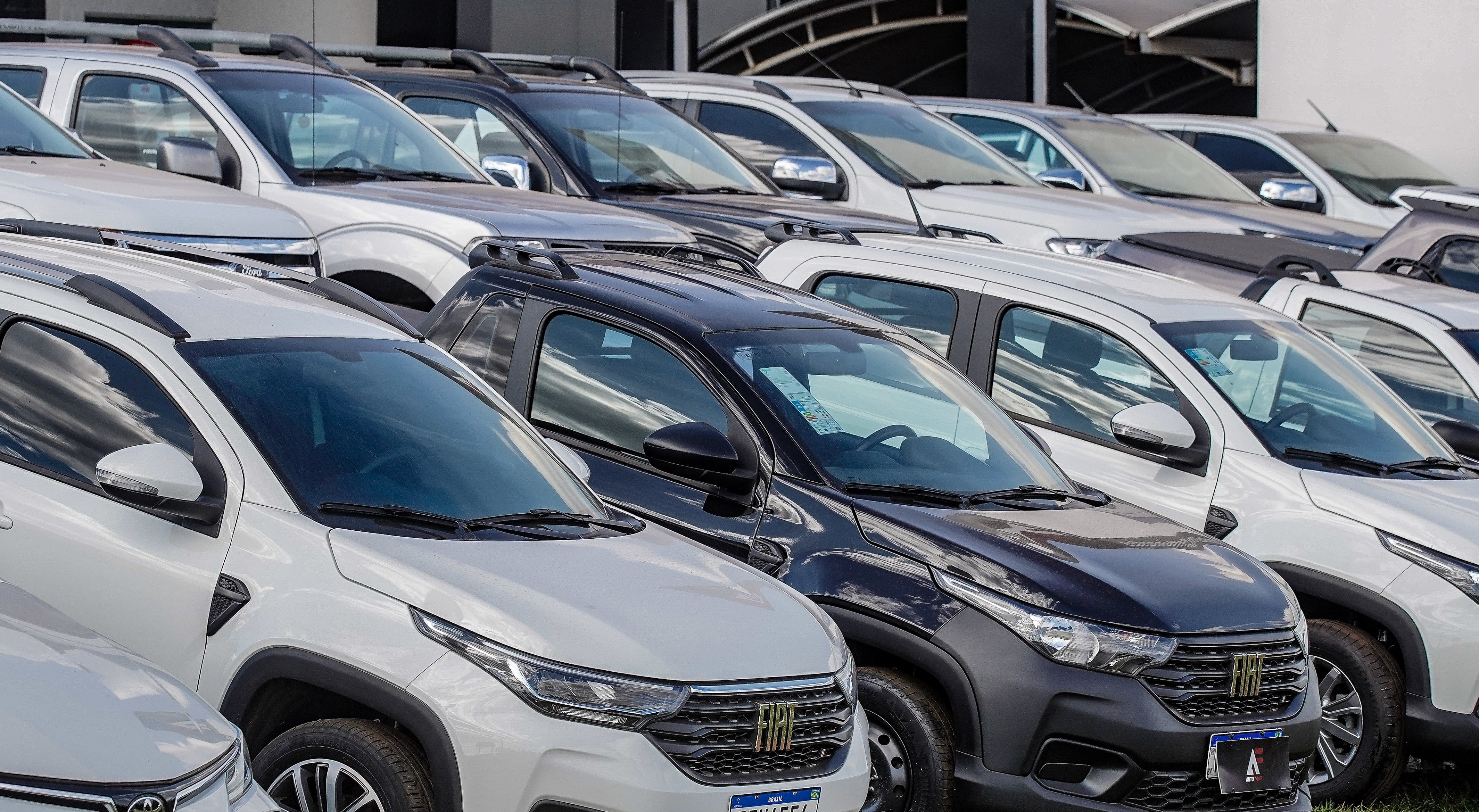 Fileira de carros novos, incluindo SUVs e picapes, estacionados ao ar livre. Há veículos brancos e um preto em destaque no centro, com detalhes em cinza escuro e rack de teto. O céu reflete nos para-brisas, sugerindo um dia claro.
